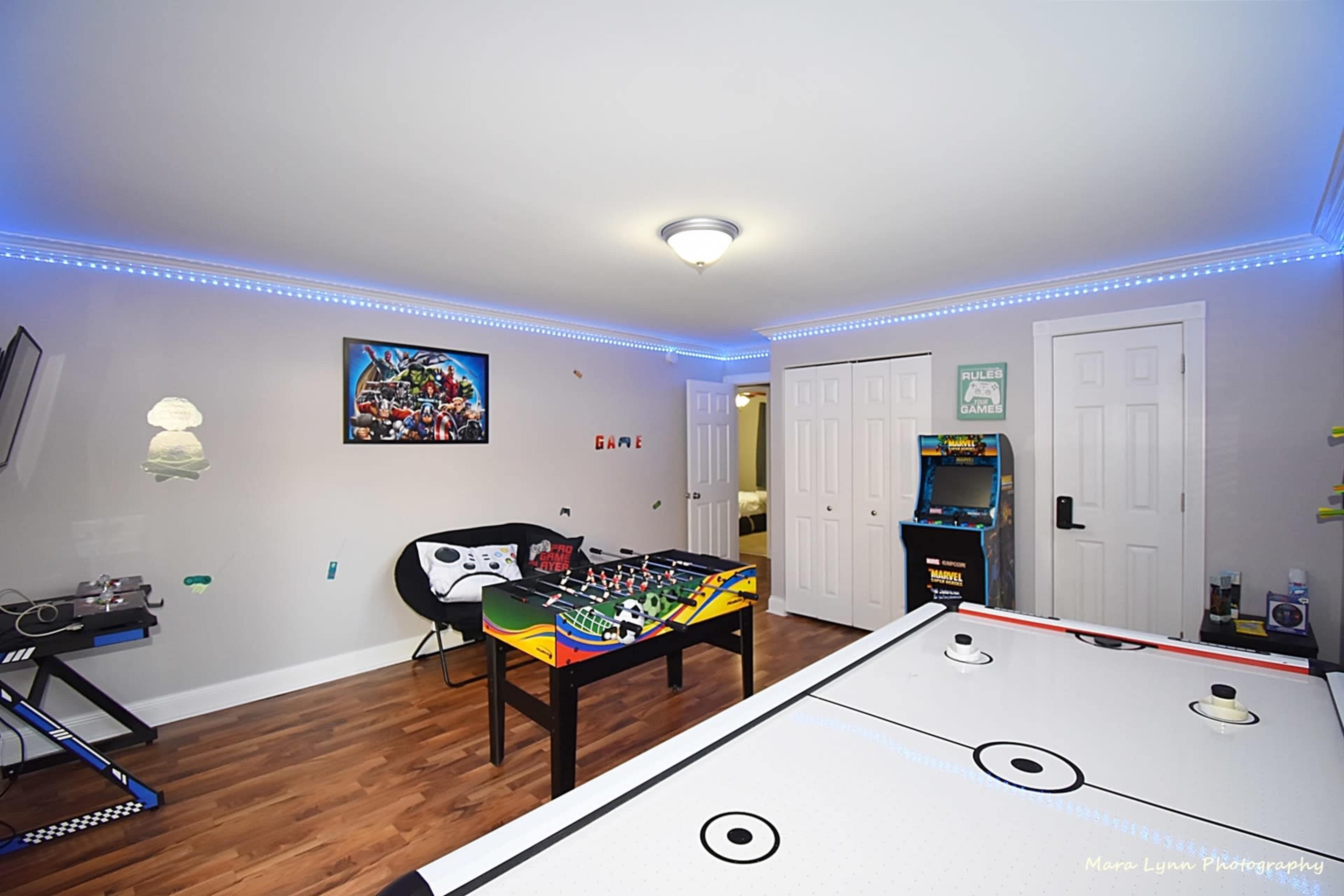 The image shows a well-lit game room featuring an air hockey table, a pinball machine, and a table with a colorful game setup, surrounded by blue LED lighting and a wall-mounted TV.