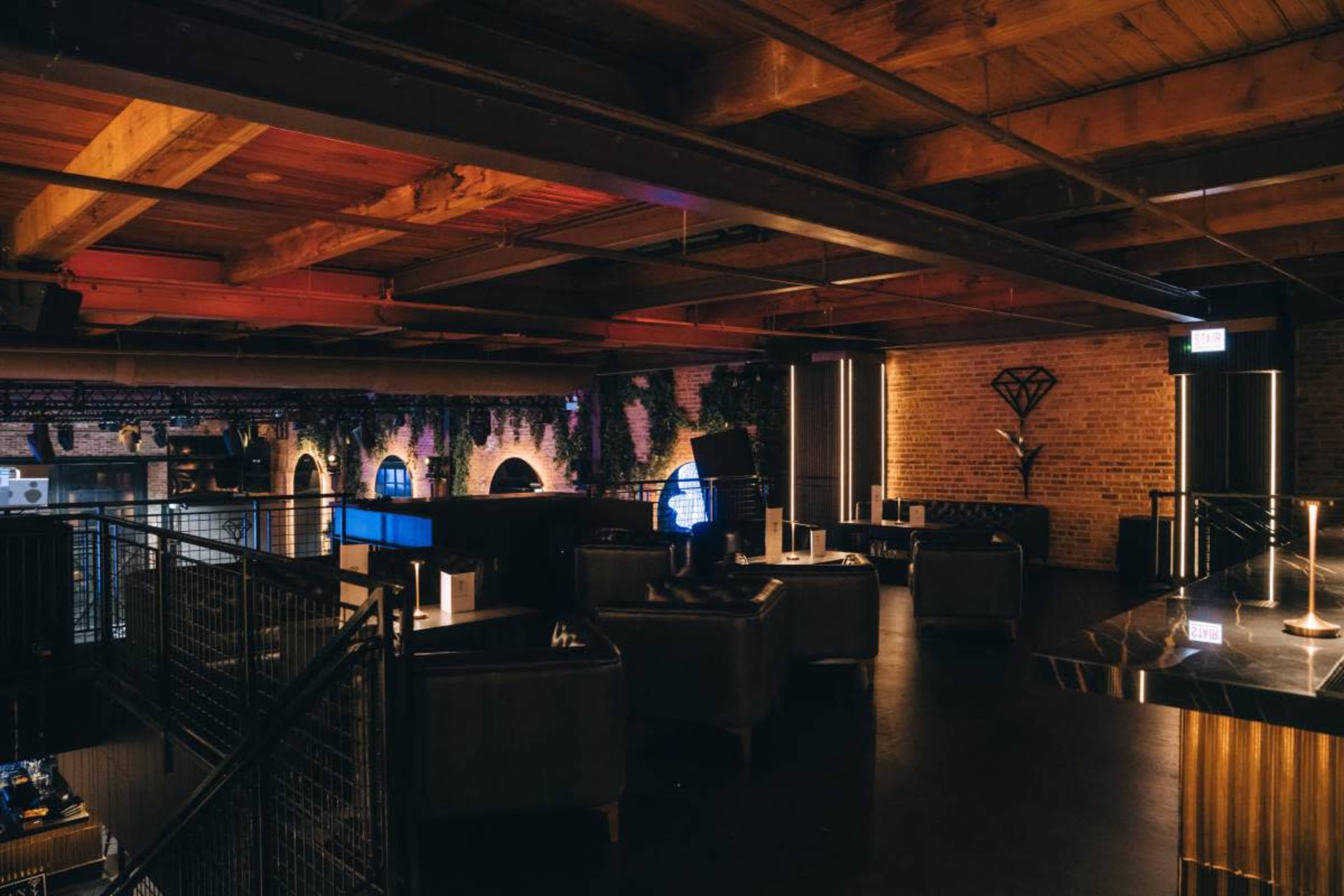 The image shows an upscale lounge with dark wood beams, leather seating, and a bar area, illuminated by soft lighting and surrounded by brick walls and greenery.