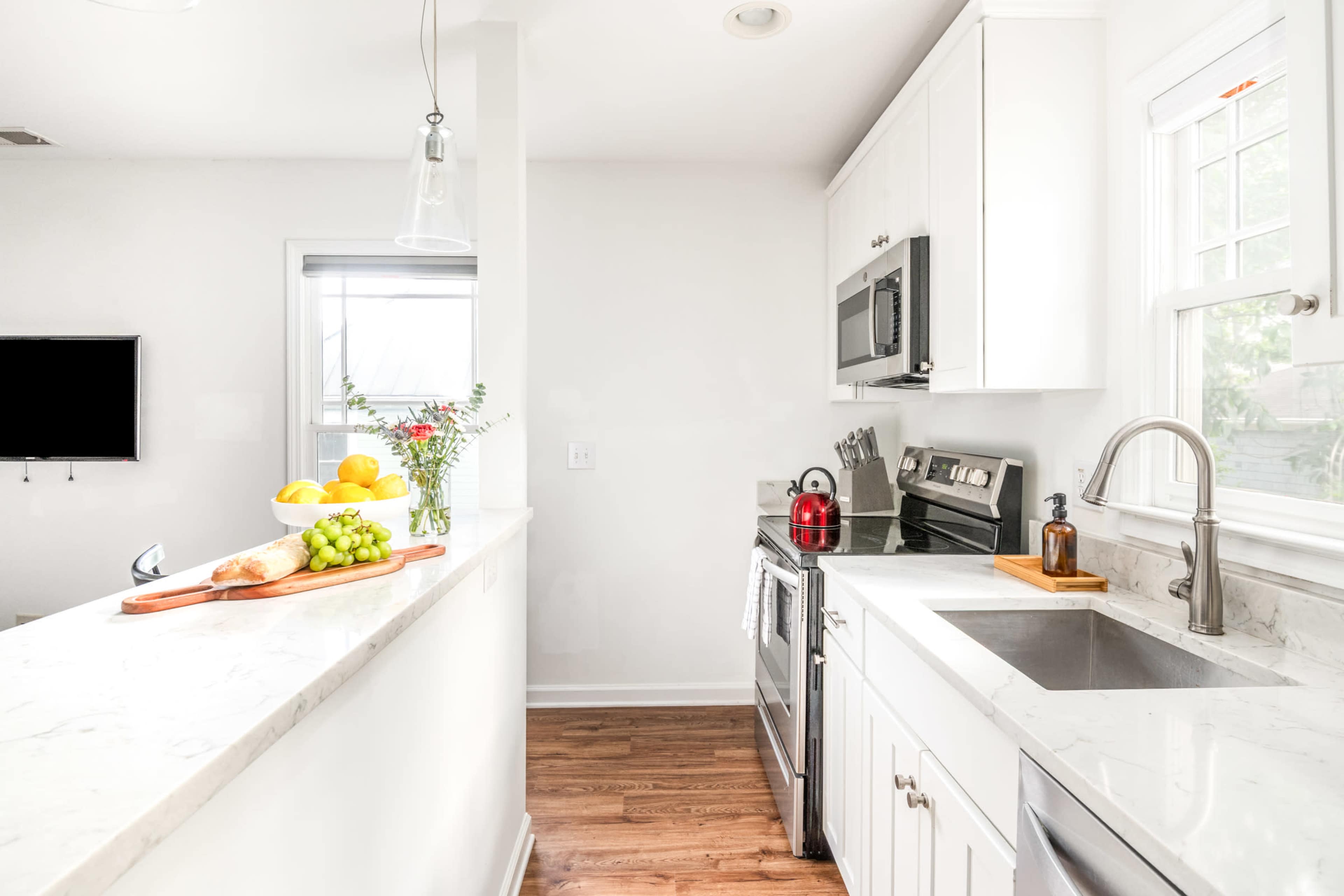The image shows a modern kitchen with white cabinetry, stainless steel appliances, a marble countertop, and a window letting in natural light.