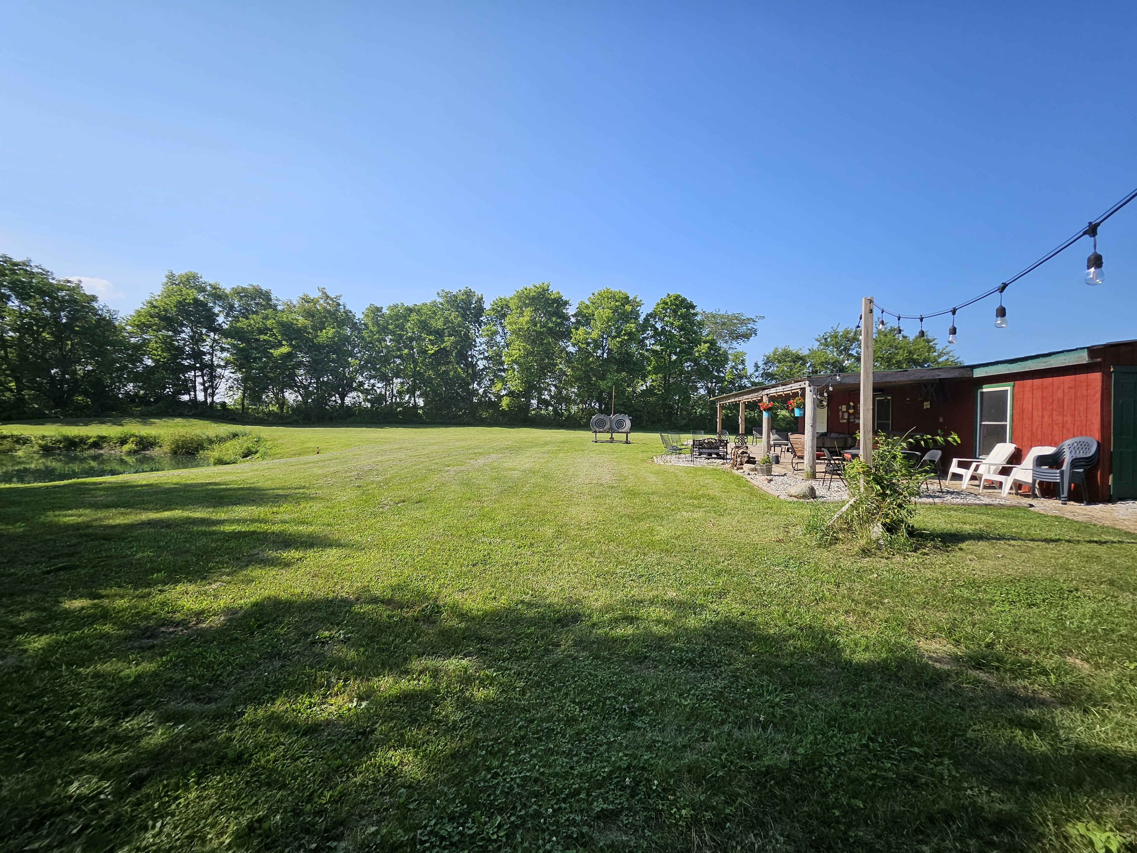 A red building with a patio overlooks a grassy field bordered by trees under a clear blue sky.