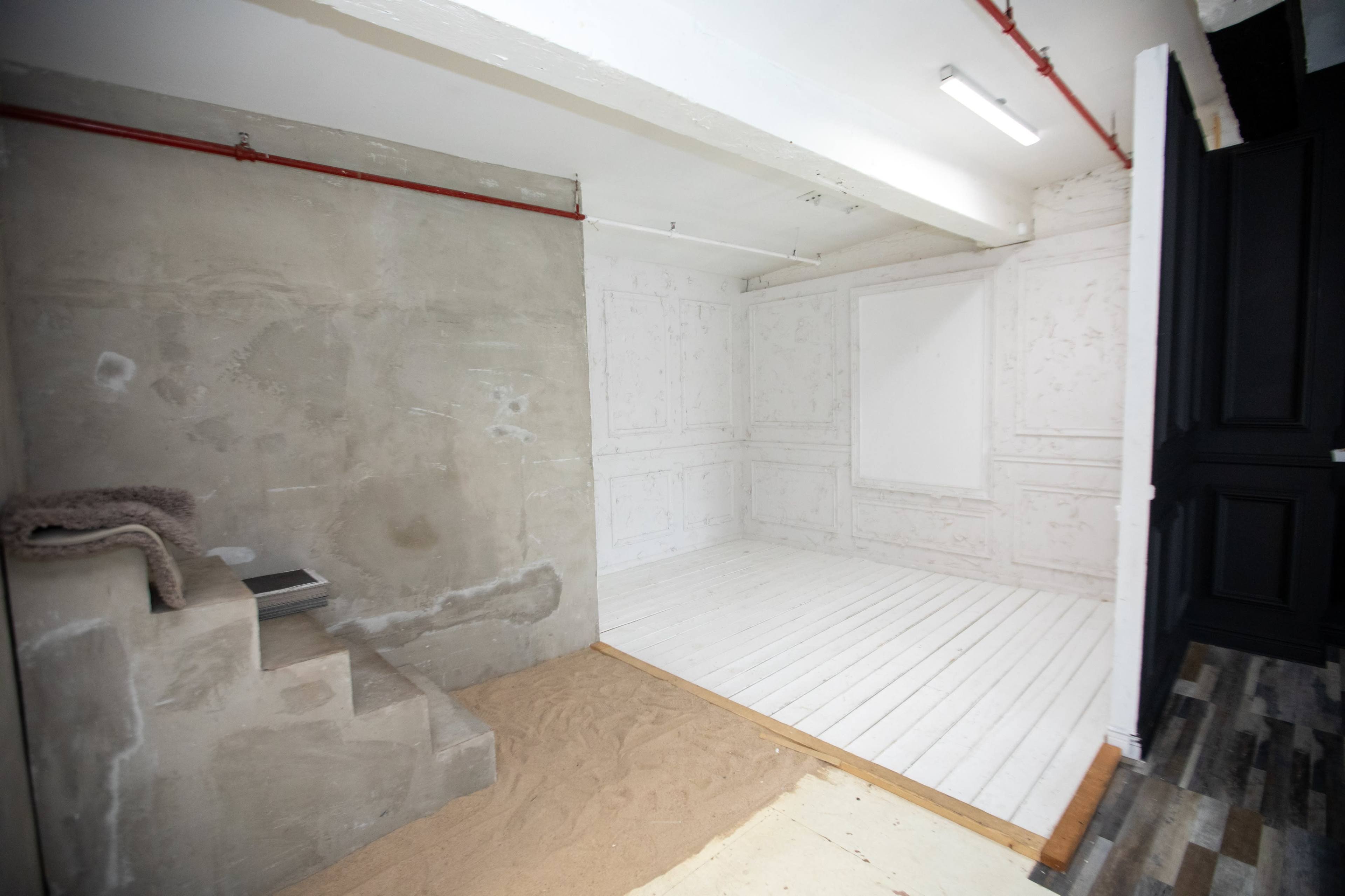 The image shows a partially constructed room with a concrete wall, white paneling, wooden flooring, and a section covered with sand.