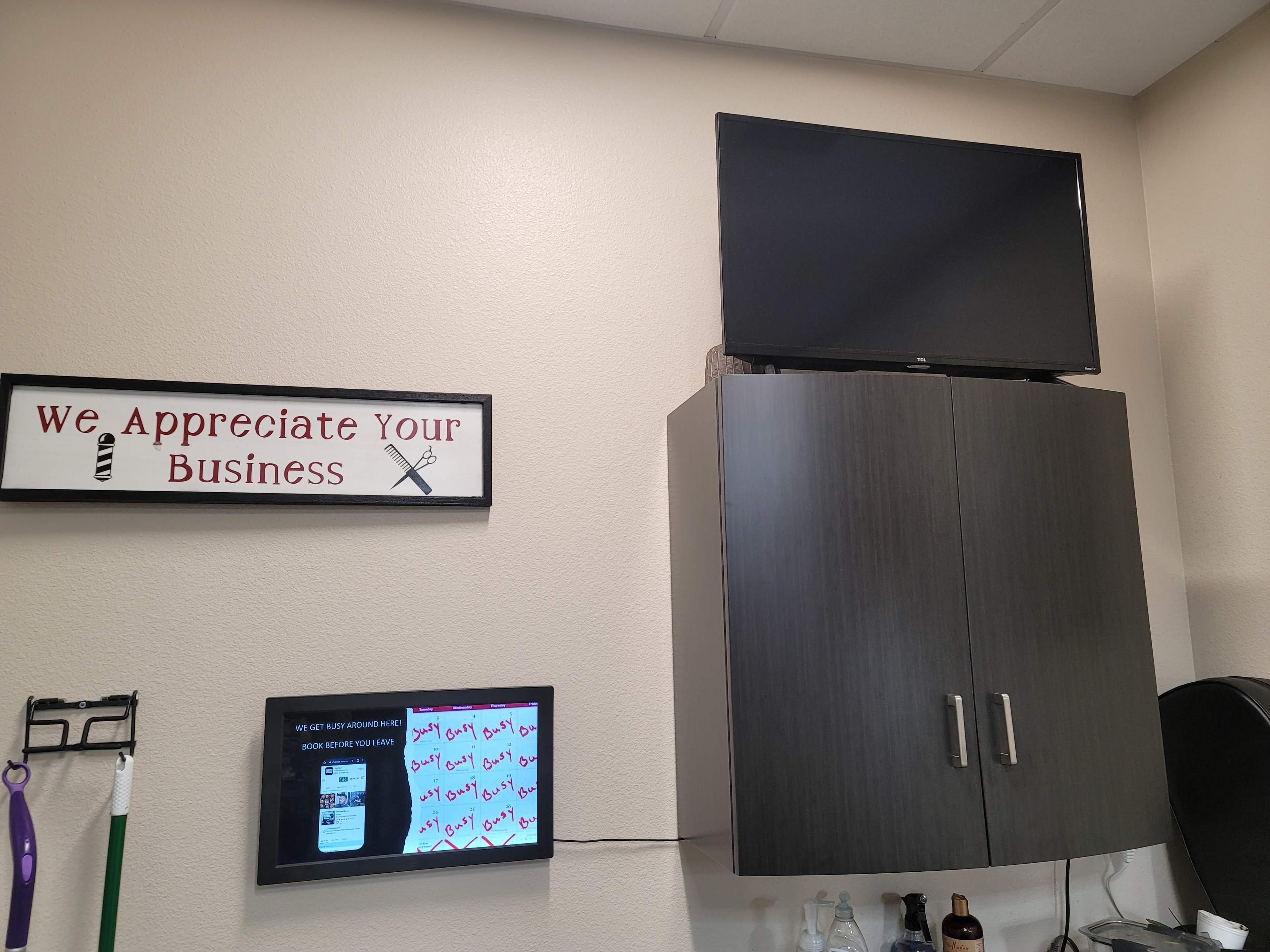 A wall-mounted television above a dark cabinet in a business setting, accompanied by a sign that reads "We Appreciate Your Business" and a digital display featuring promotional content.