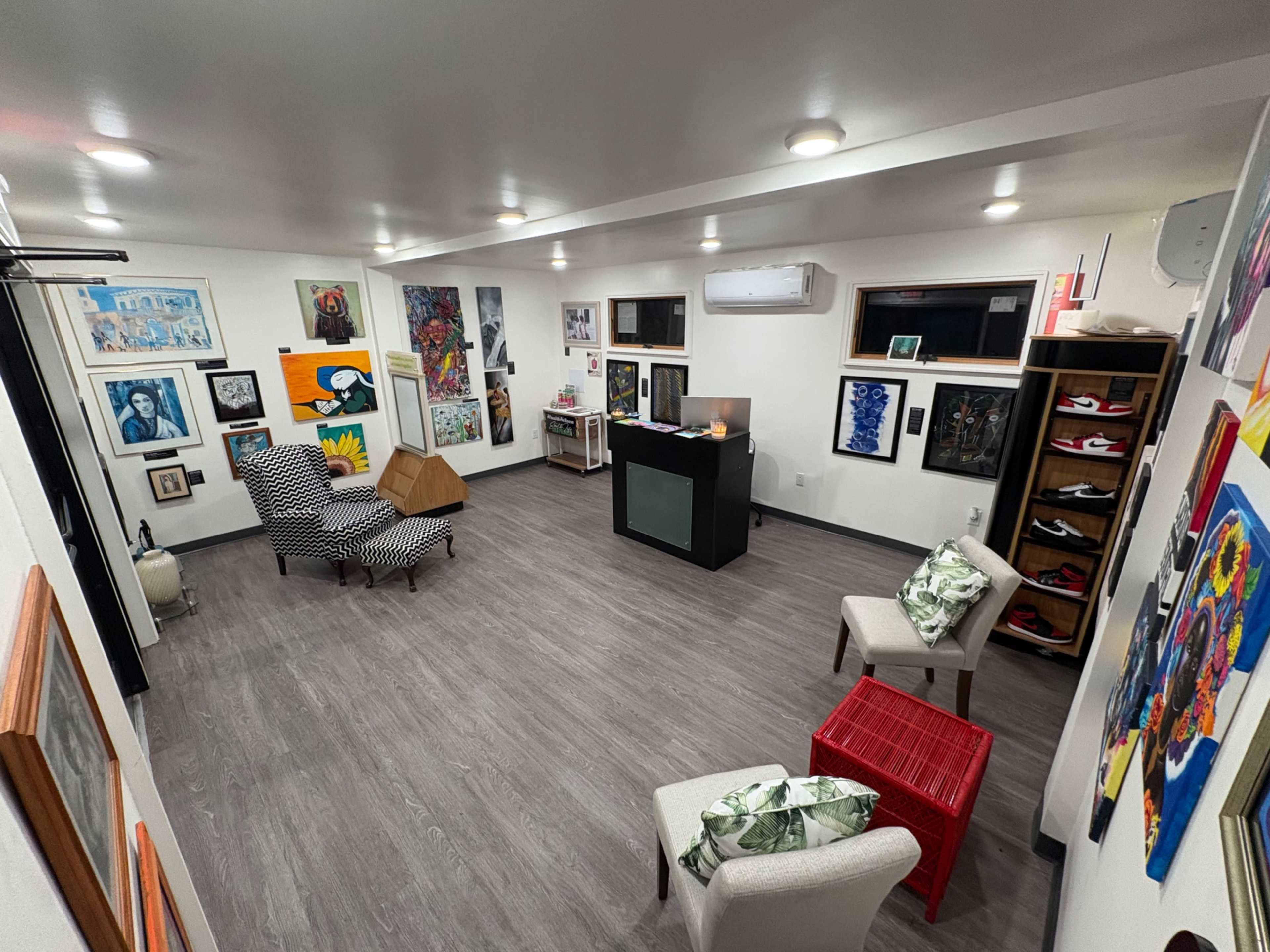 The image shows an art-filled room with various artworks on the walls, a small reception area with furniture, and a wooden floor.