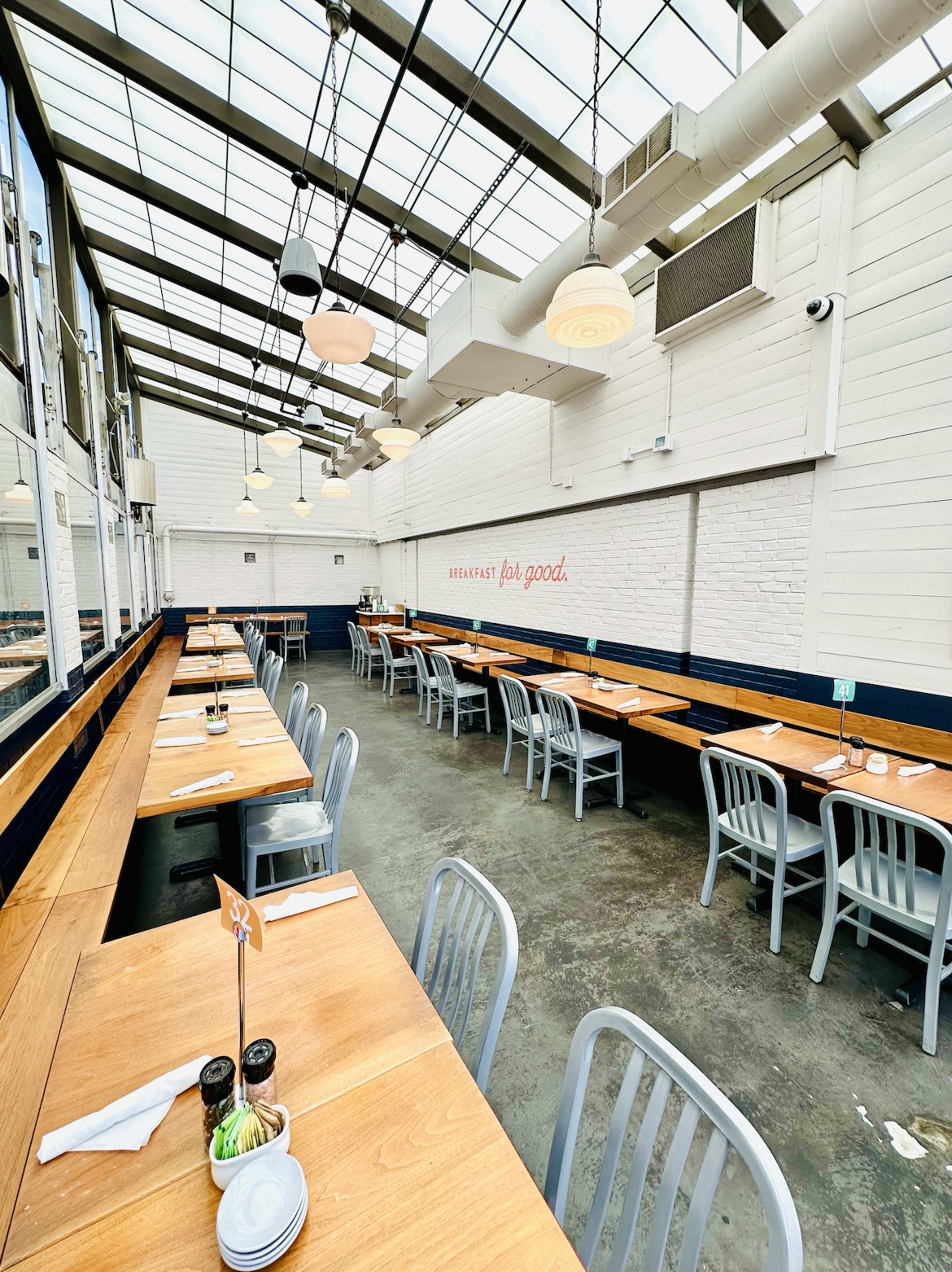 The image shows a spacious restaurant interior with long wooden tables and metal chairs arranged under a glass ceiling.