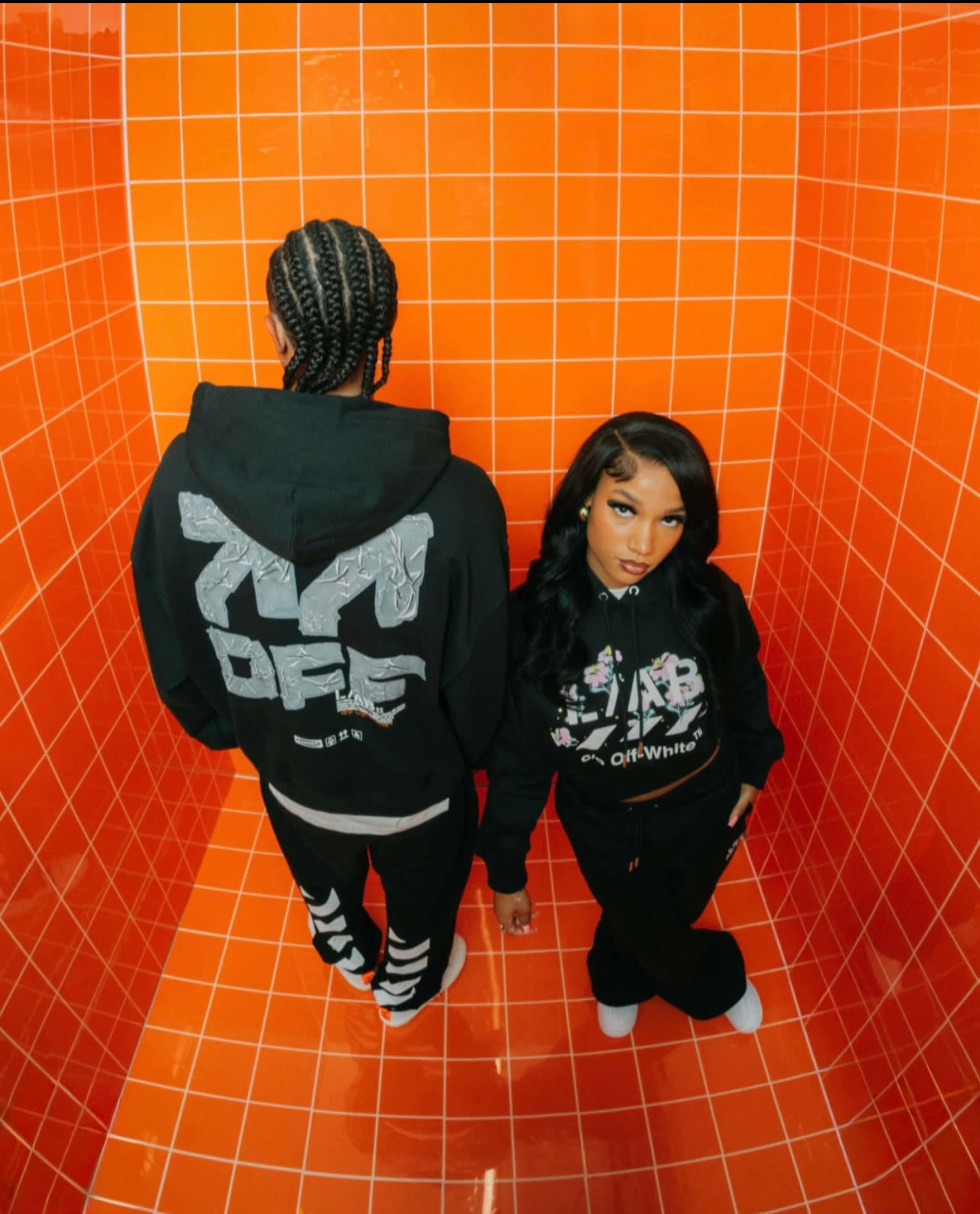 Two individuals stand in a brightly colored orange tiled room, wearing fashionable streetwear hoodies.