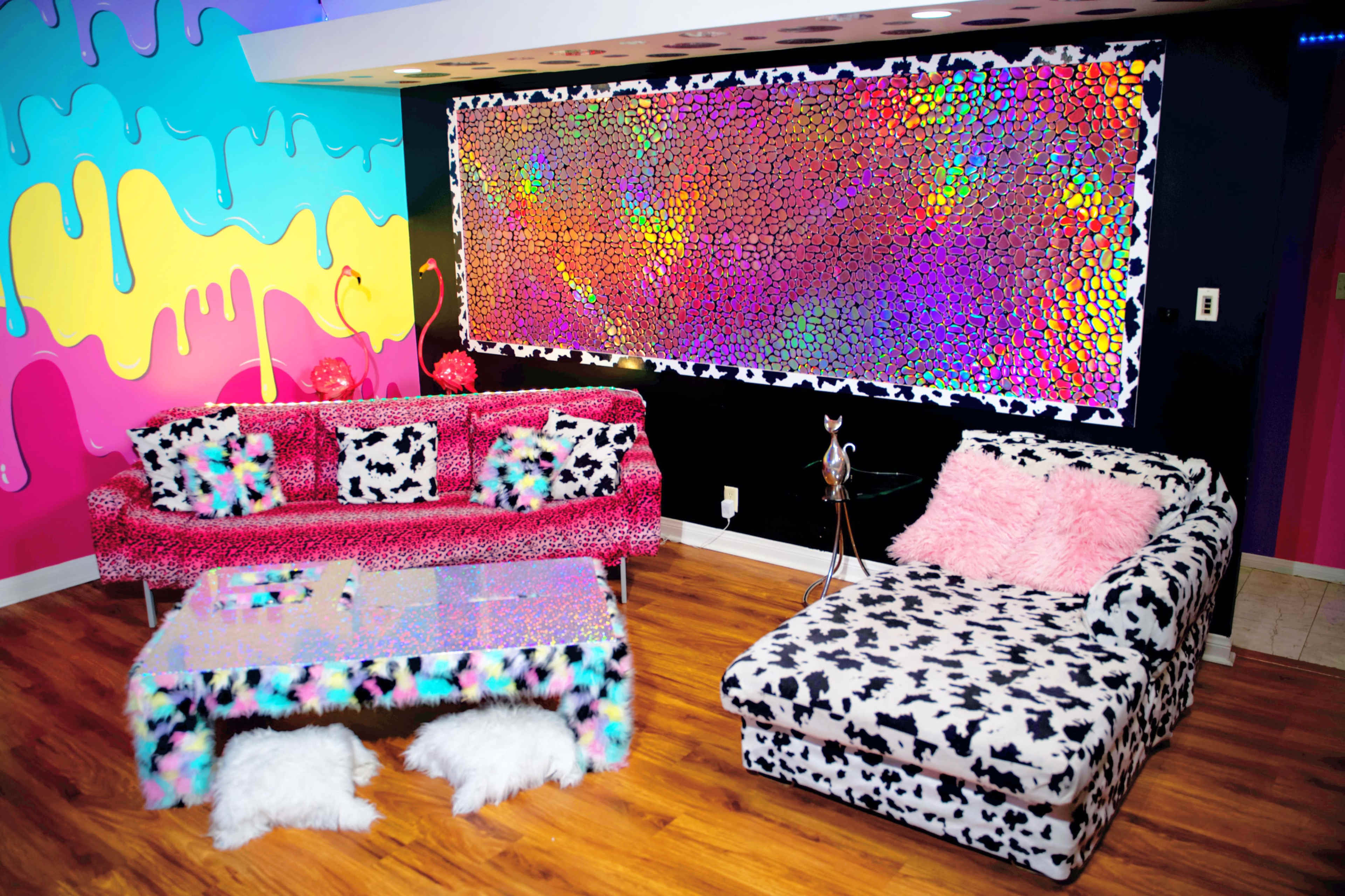 A brightly colored room features a pink and blue wall with a multicolored abstract art piece, matched with animal-print furniture and a sparkly glass coffee table.