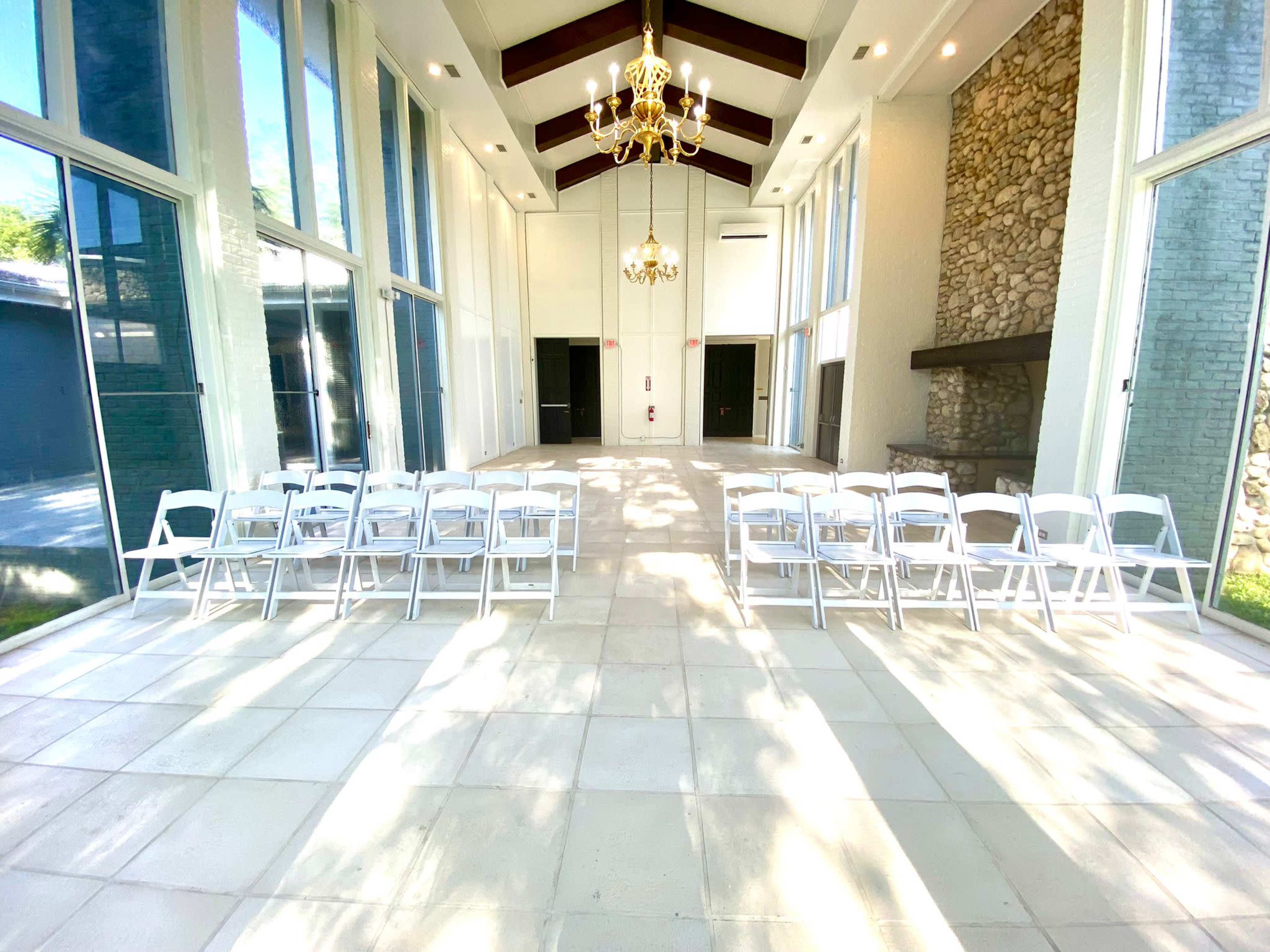 The image shows a spacious indoor area with large windows, featuring rows of white folding chairs arranged for an event, and a stone fireplace against one wall.