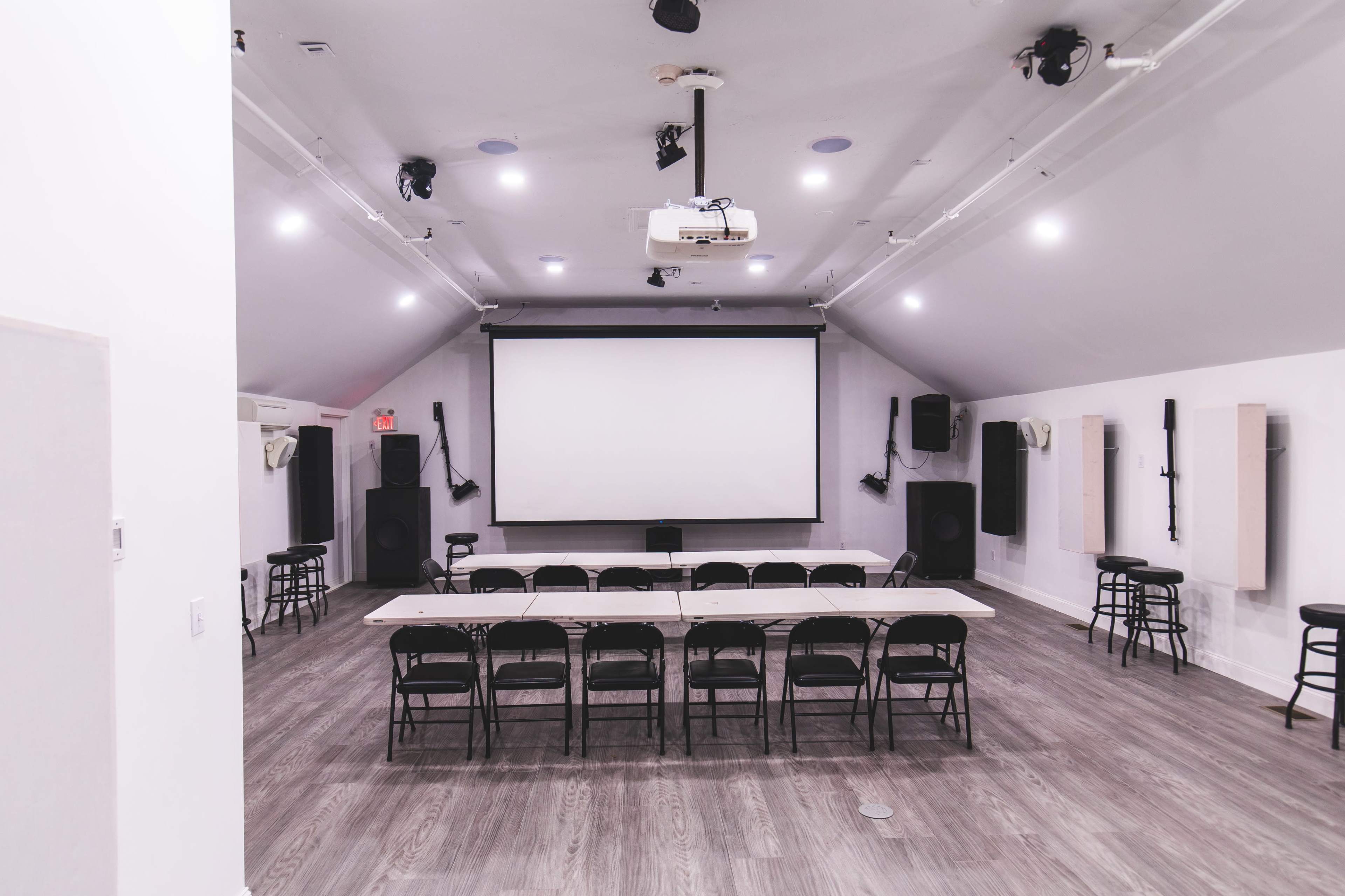 The image shows a spacious presentation room with a large screen, surround sound speakers, and a long table set with black chairs arranged in a semi-circle.