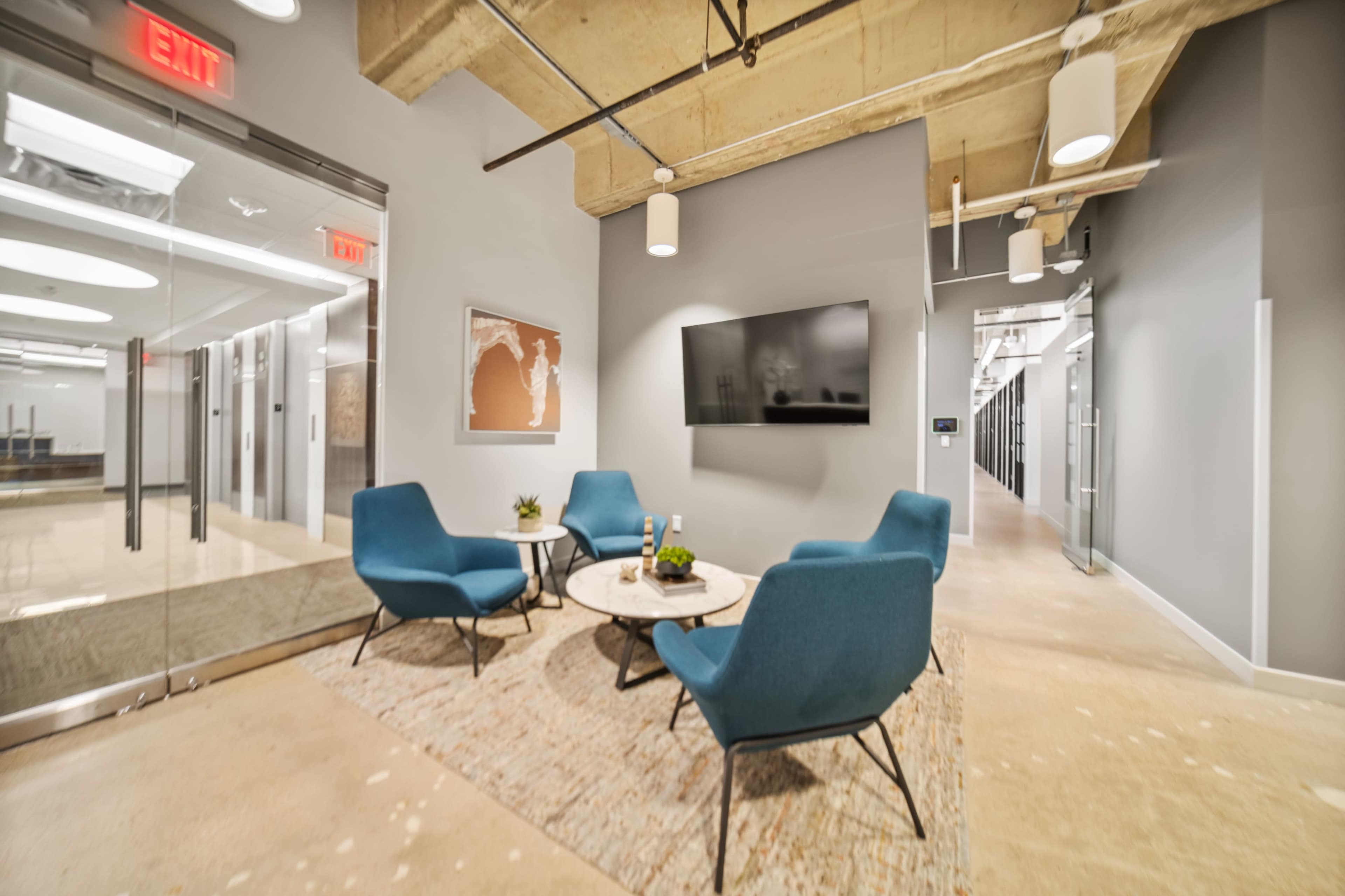 The image shows a modern office space featuring a seating area with blue chairs, a small coffee table, and a large wall-mounted television, adjacent to a corridor leading to other office rooms.
