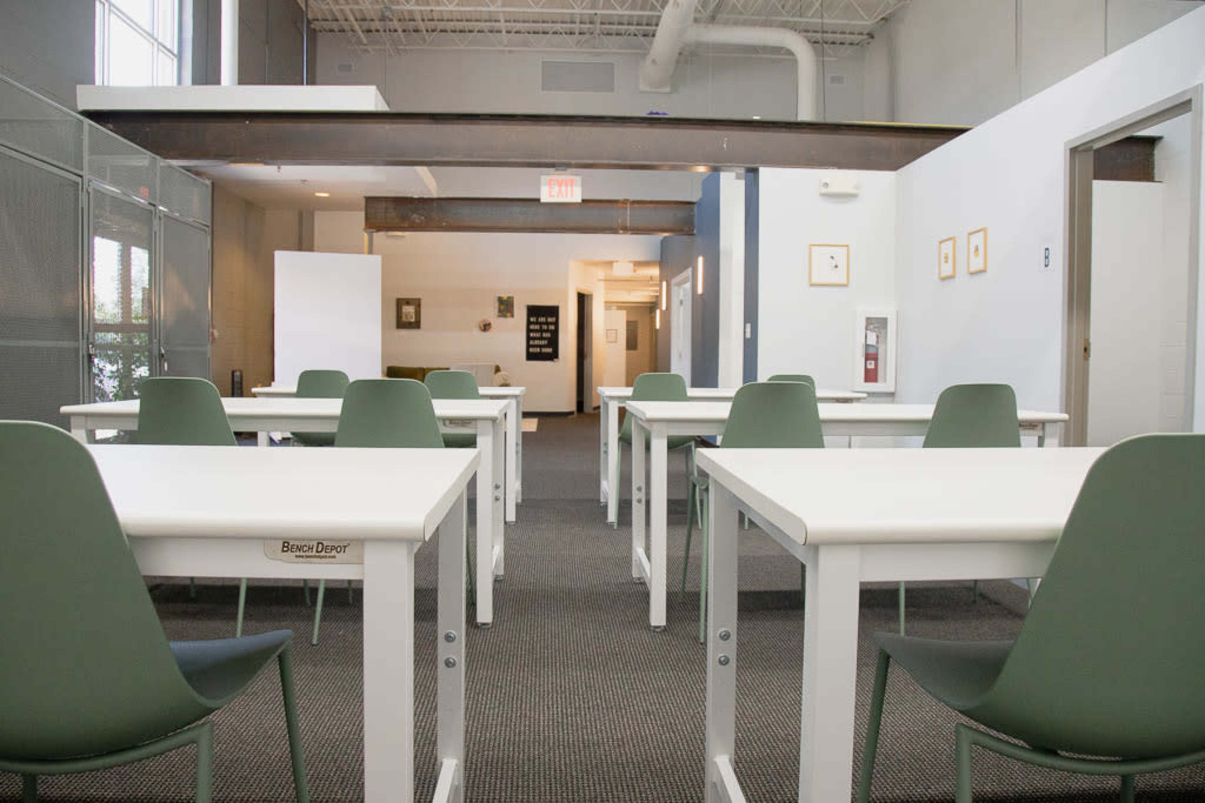 A classroom with several white tables and green chairs arranged in neat rows, facing a doorway at the end of the room.
