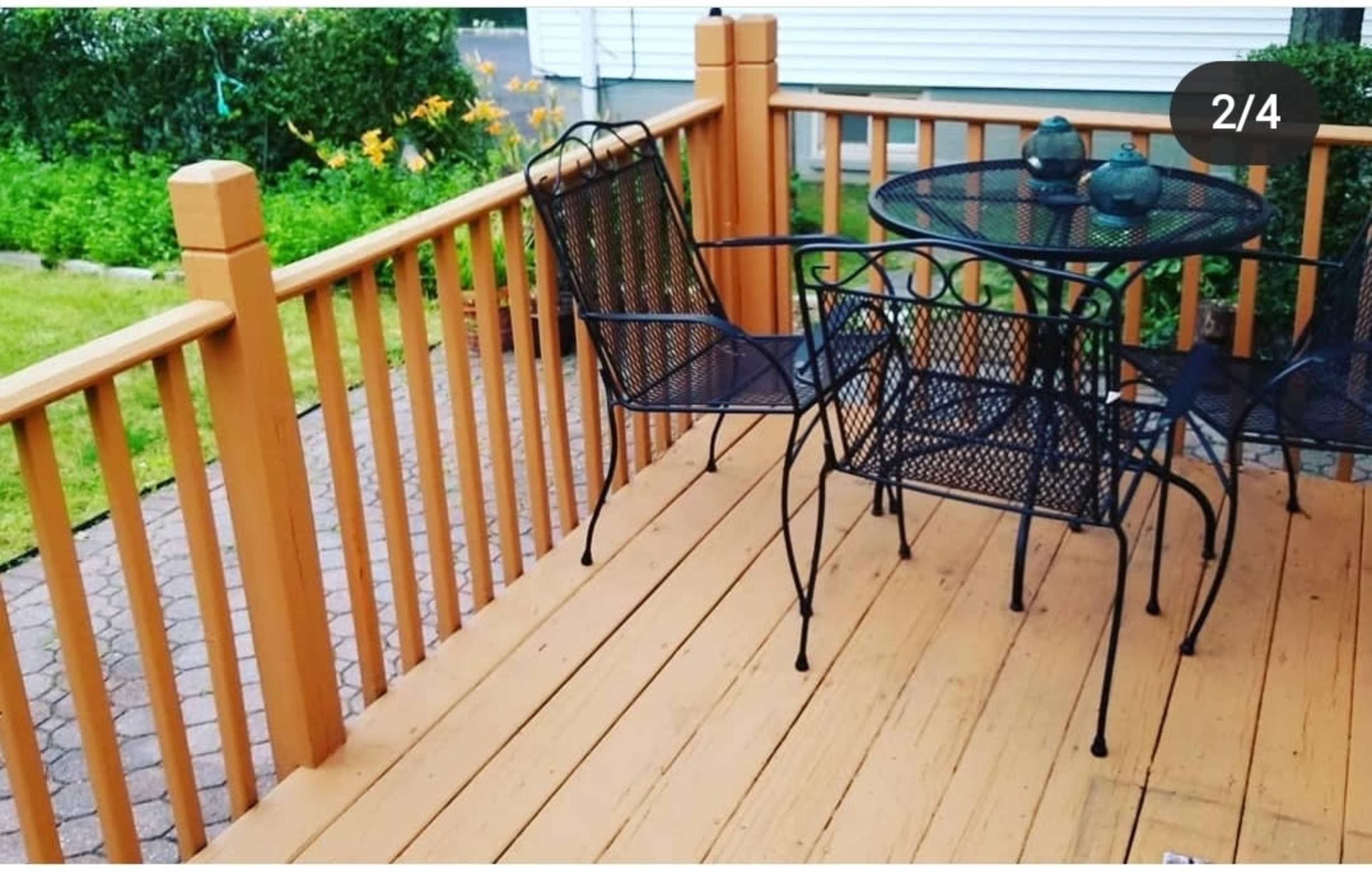 A wooden deck with a black metal table and two chairs set on it, surrounded by green plants in the background.