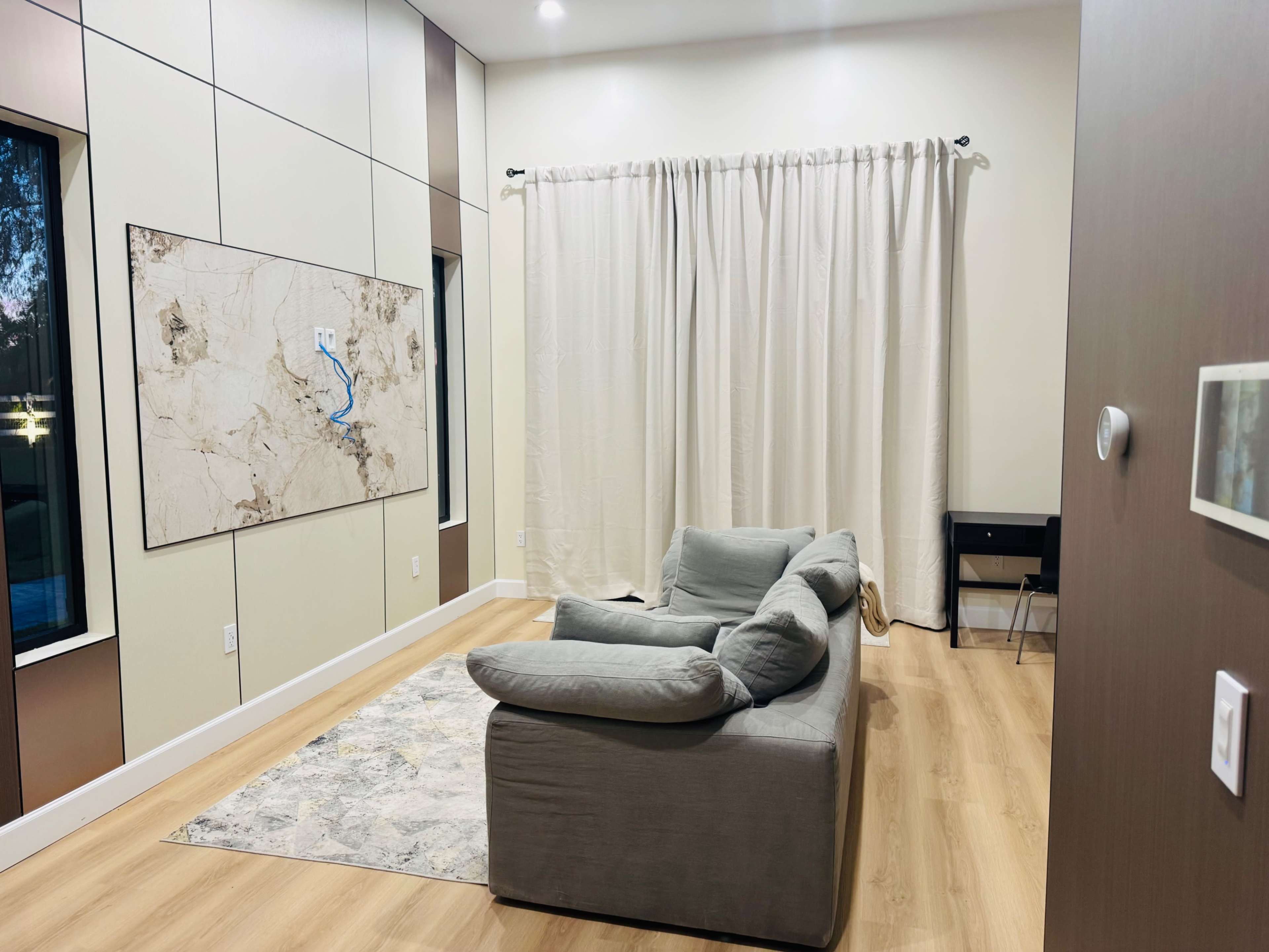 The image shows a modern living room with a gray sectional sofa, a light area rug, a large wall art piece, and a set of windows beside a curtain.