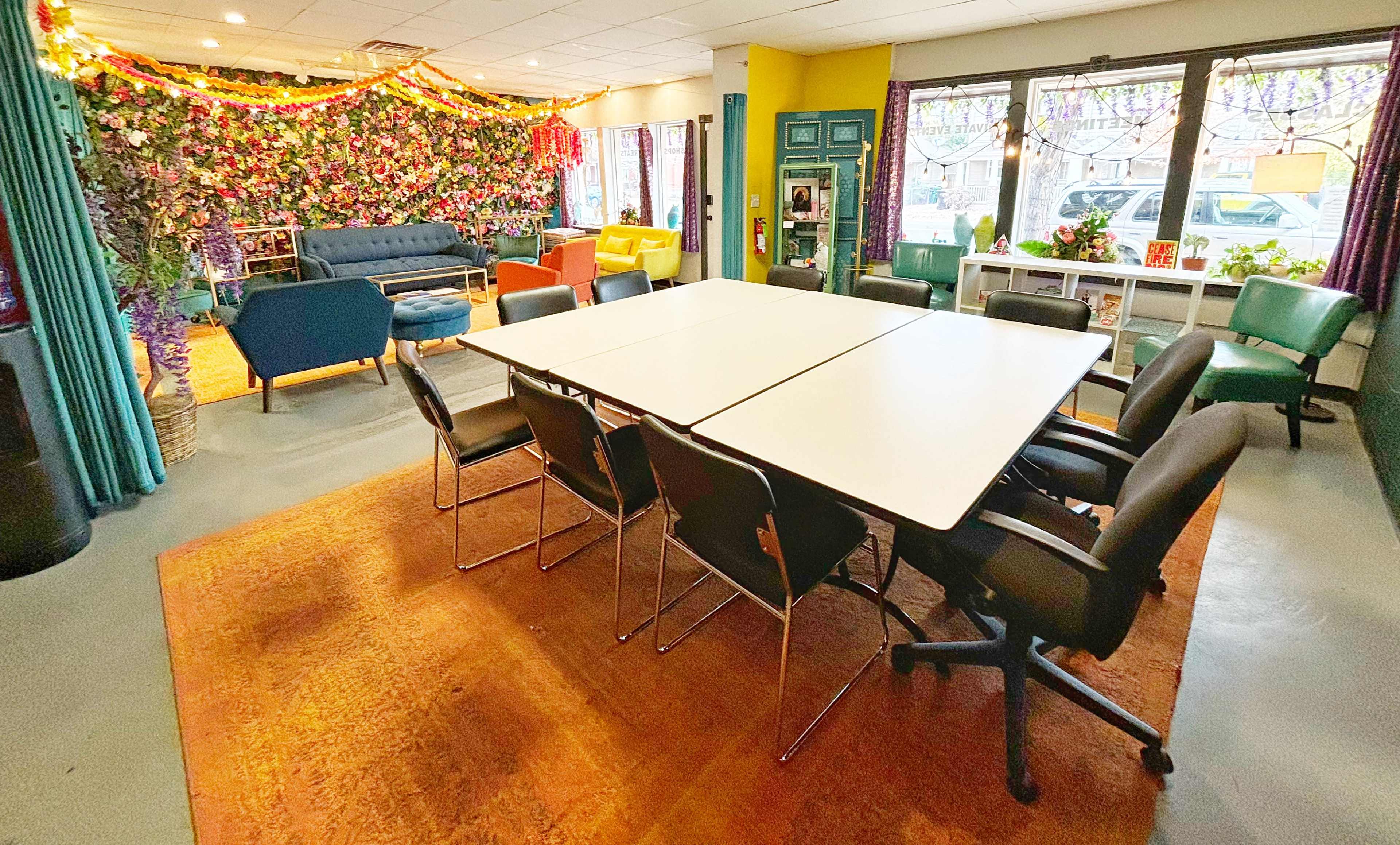 The image shows a spacious meeting room featuring a large white table surrounded by black chairs, with colorful decorative elements and a wall of greenery in the background.