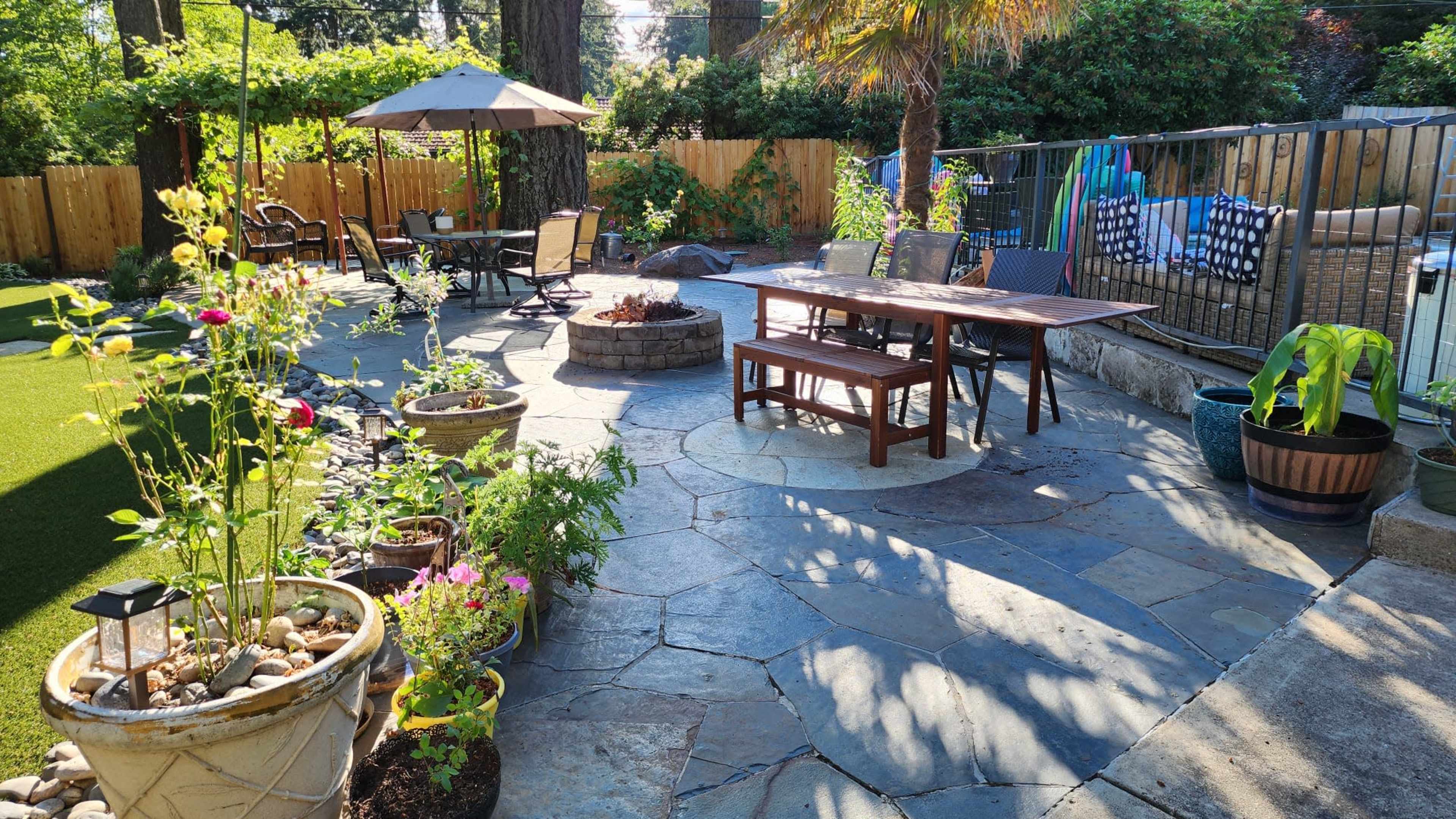 A backyard patio features a stone surface with a table and chairs, surrounded by potted plants and a fire pit, with a pool and umbrellas in the background.