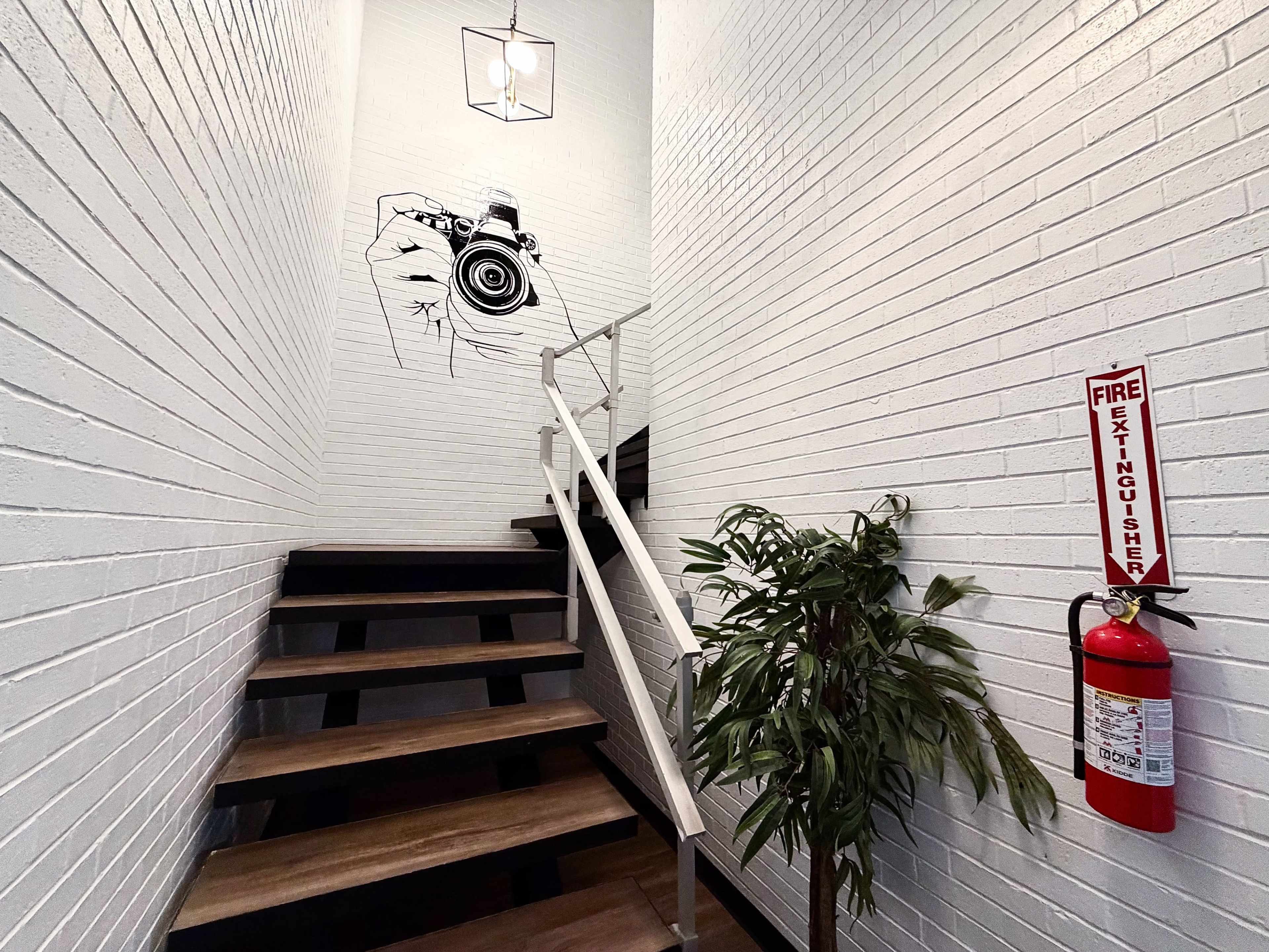 The image shows a staircase leading upward, with a light fixture hanging above, a mural of a hand holding a camera on the wall, a potted plant at the base, and a fire extinguisher mounted on the wall.