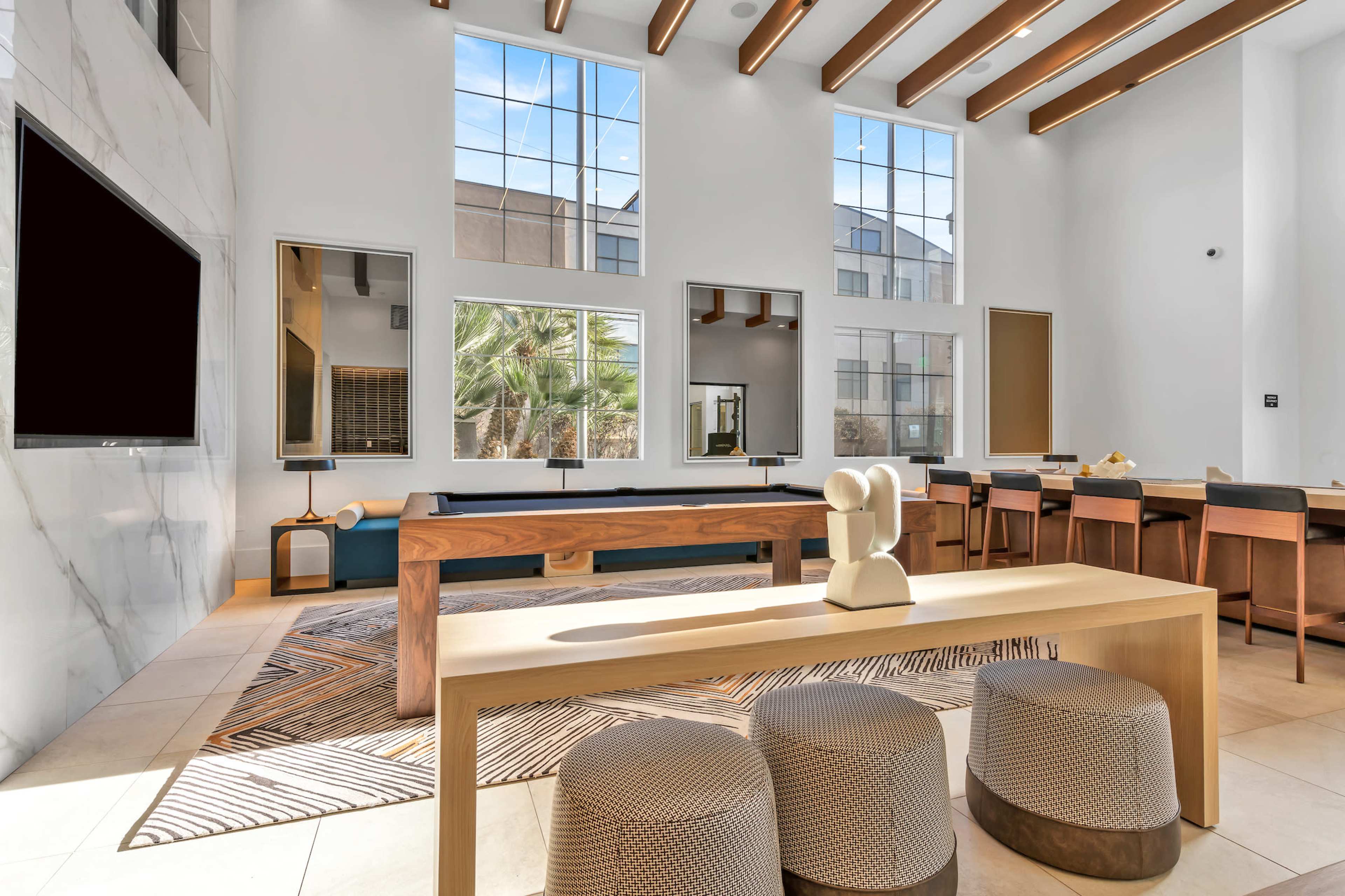 A modern living space features large windows, wooden beams, a sectional sofa, and a minimalist coffee table surrounded by chairs.