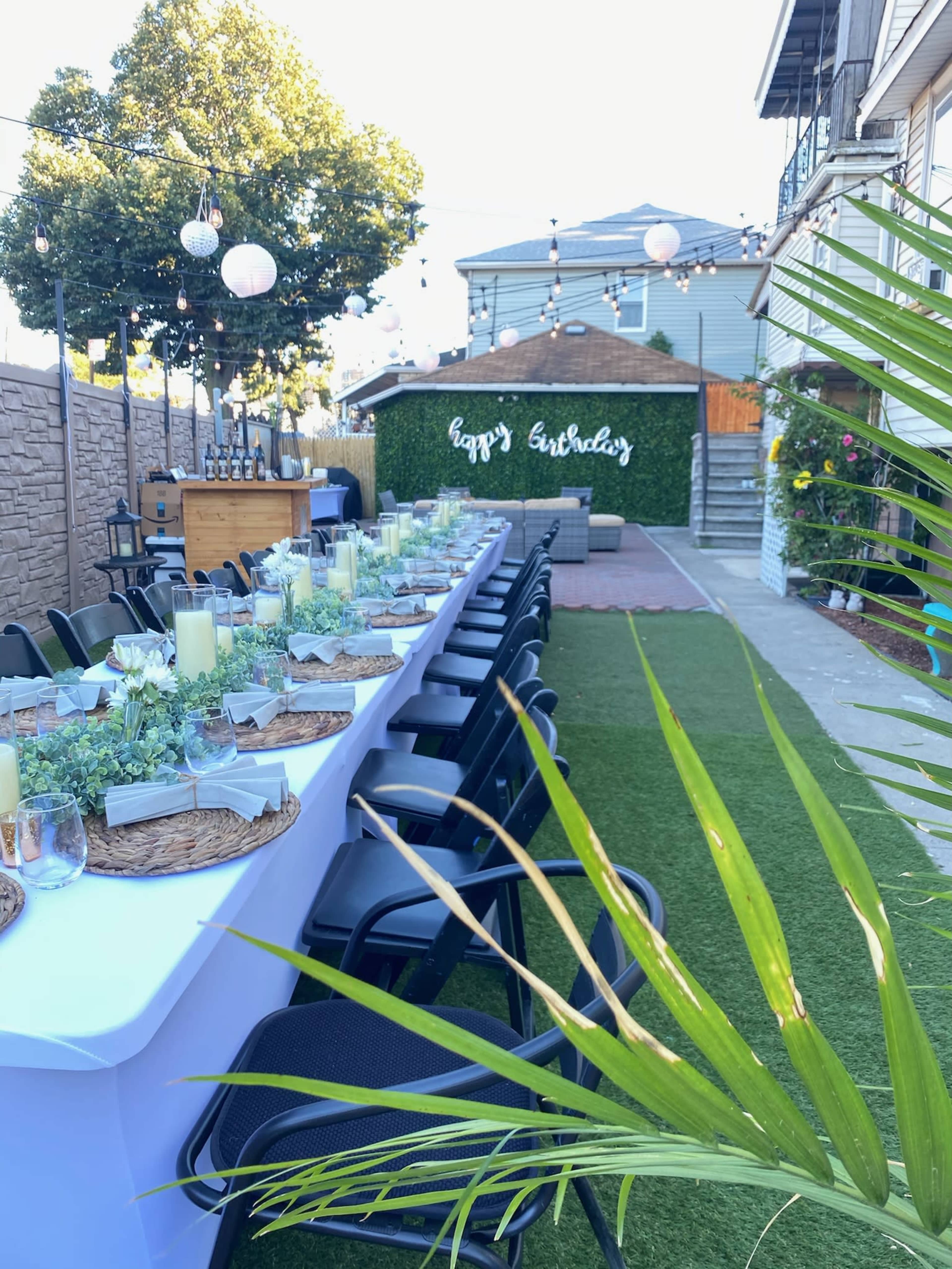 A long dining table is set with greenery and candles in a backyard decorated for a birthday celebration, featuring a "Happy Birthday" sign and string lights overhead.