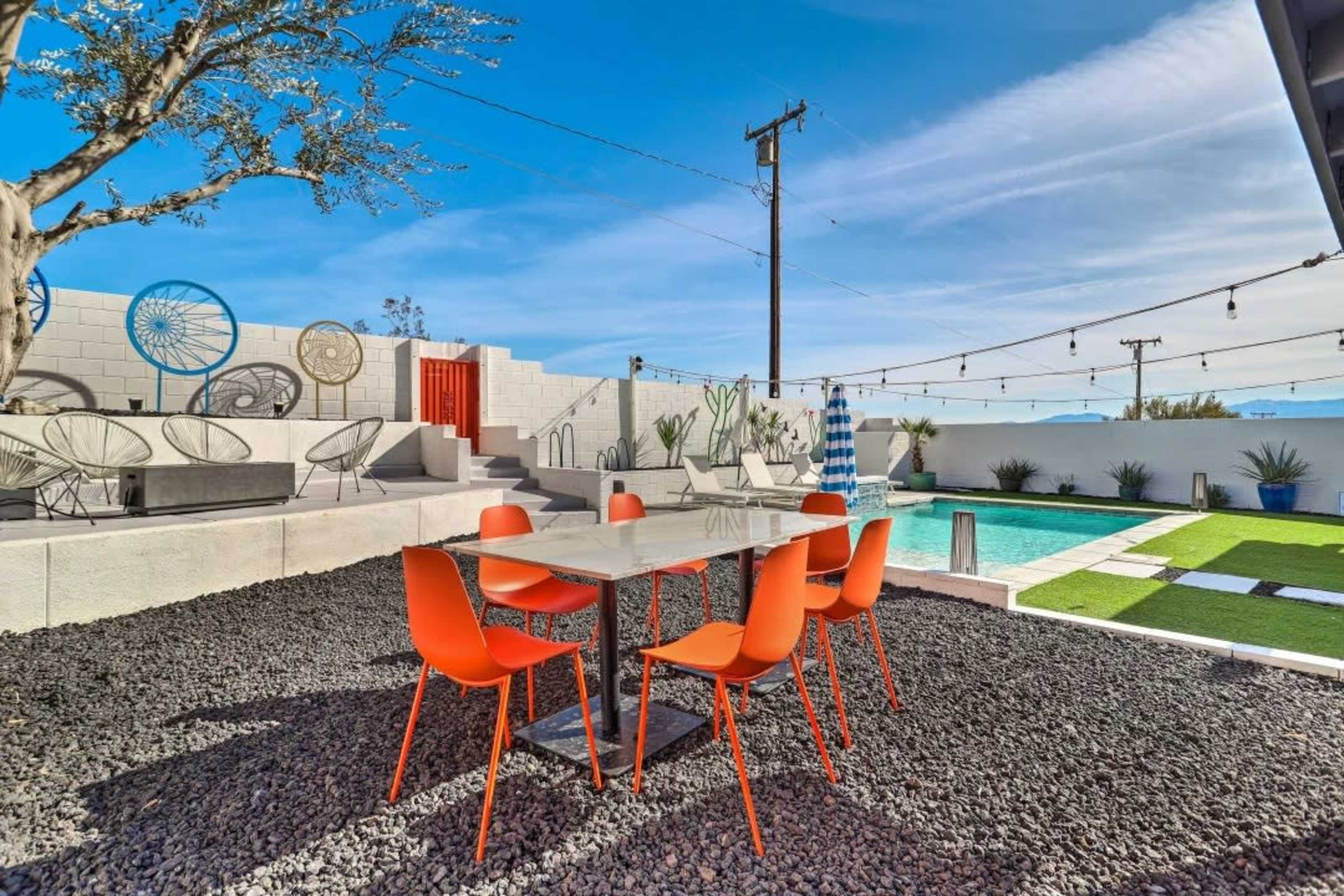 A modern outdoor dining area features a glass table surrounded by orange chairs, with a pool and landscaped stones in the background.