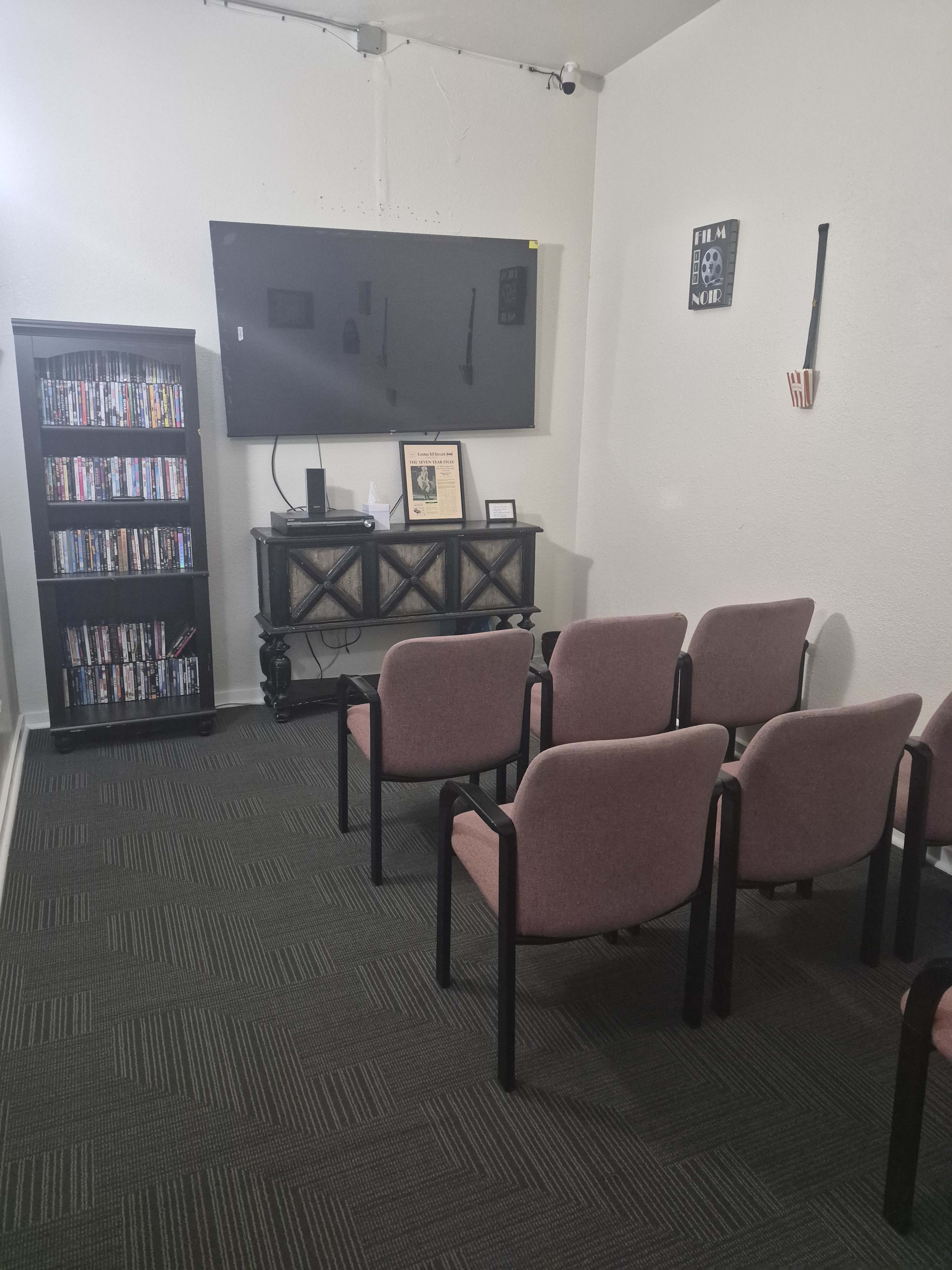 The room features a small seating area with several chairs facing a mounted television and a wooden cabinet displaying DVDs, along with a framed picture on the wall.