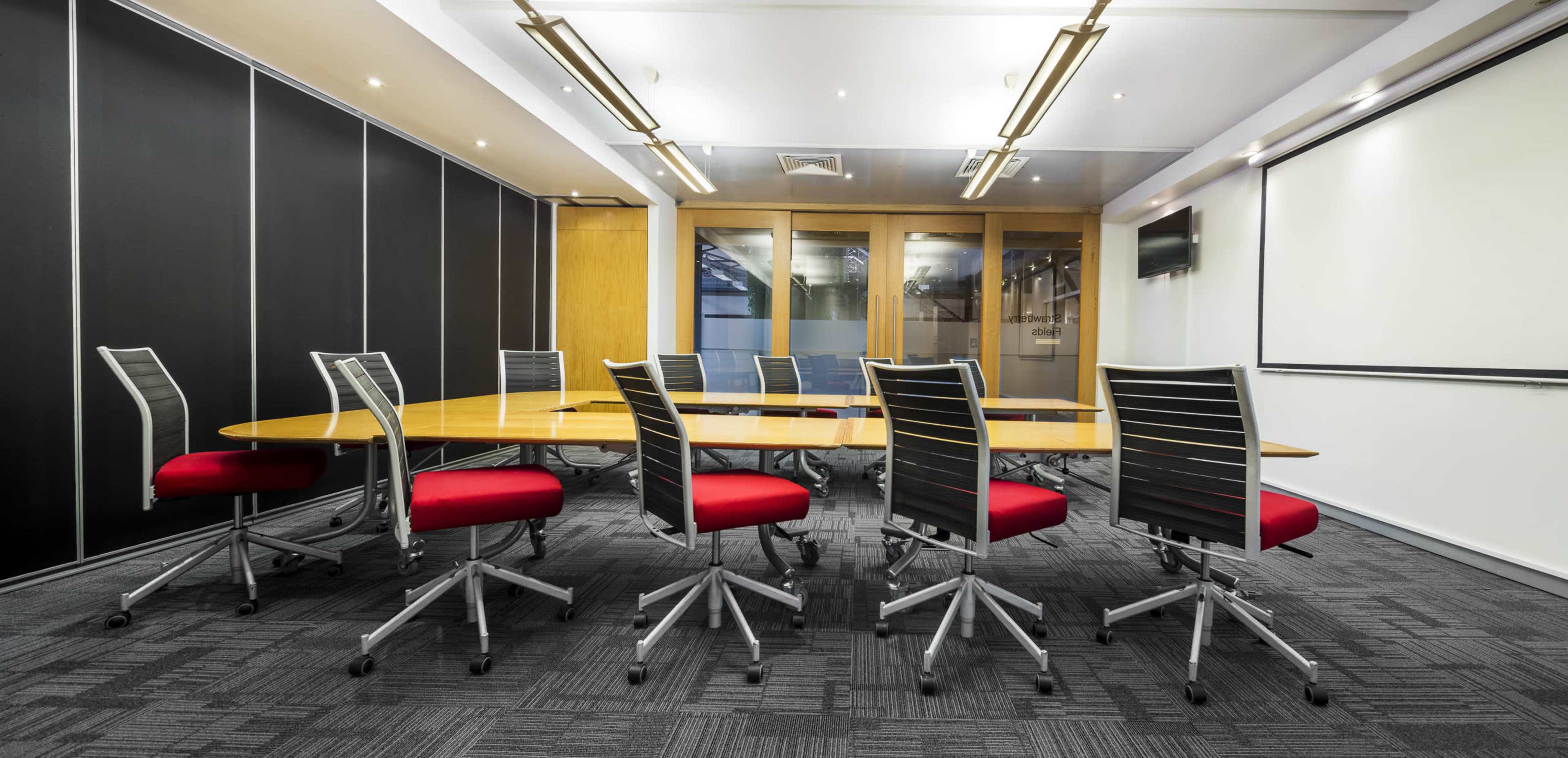 A modern conference room features a large oval table surrounded by ergonomic red office chairs and equipped with a presentation screen.