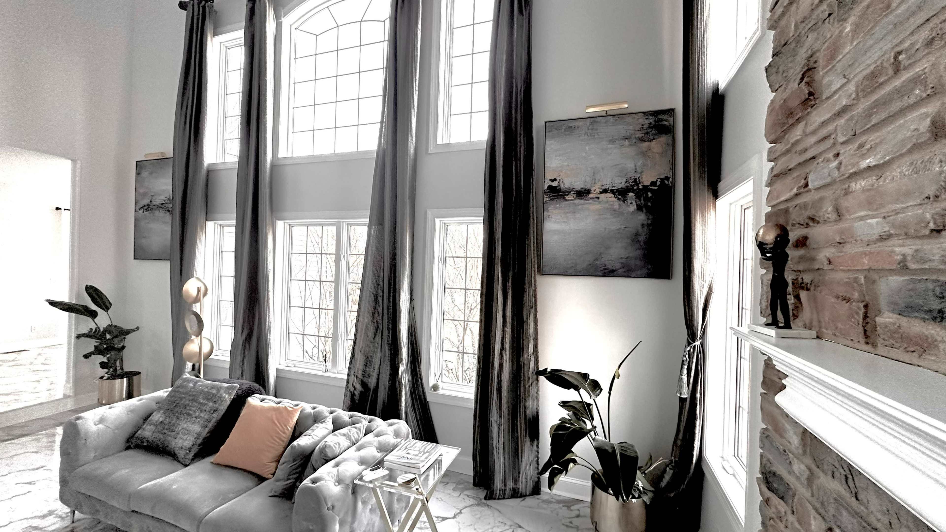 A modern living room with large windows, tall curtains, and a gray sofa adorned with a pillow, accompanied by plants and artwork on the walls.