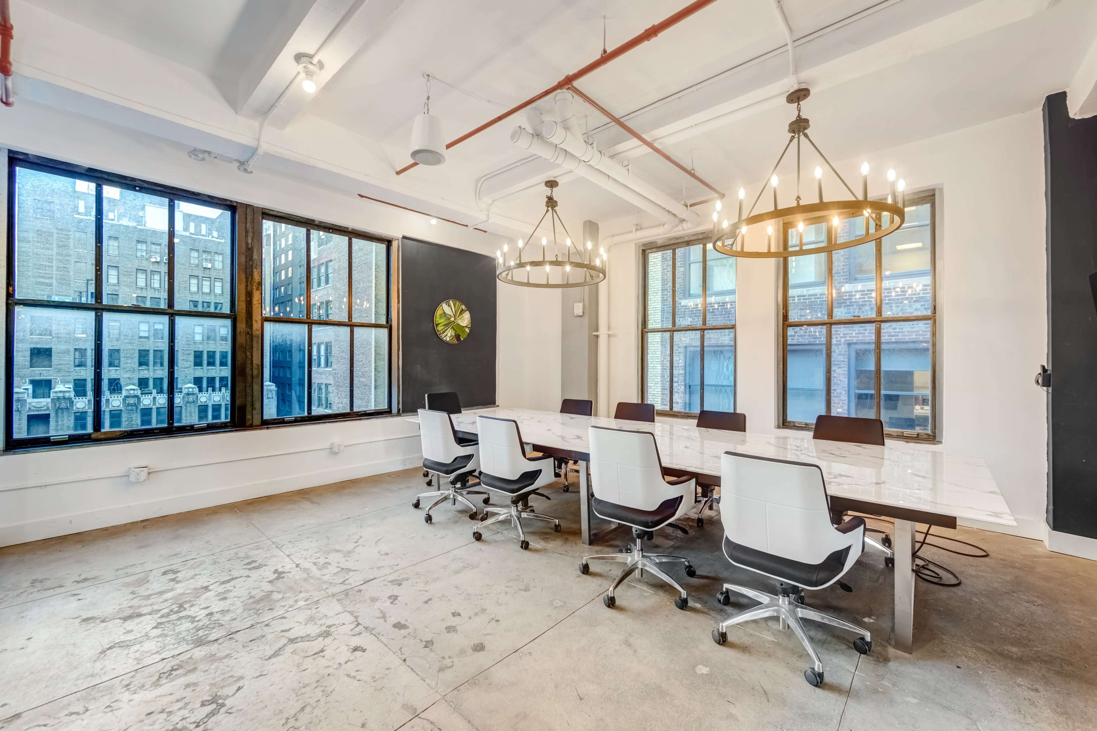 A large conference room features a long table surrounded by ergonomic chairs, with two large windows showcasing a view of city buildings.