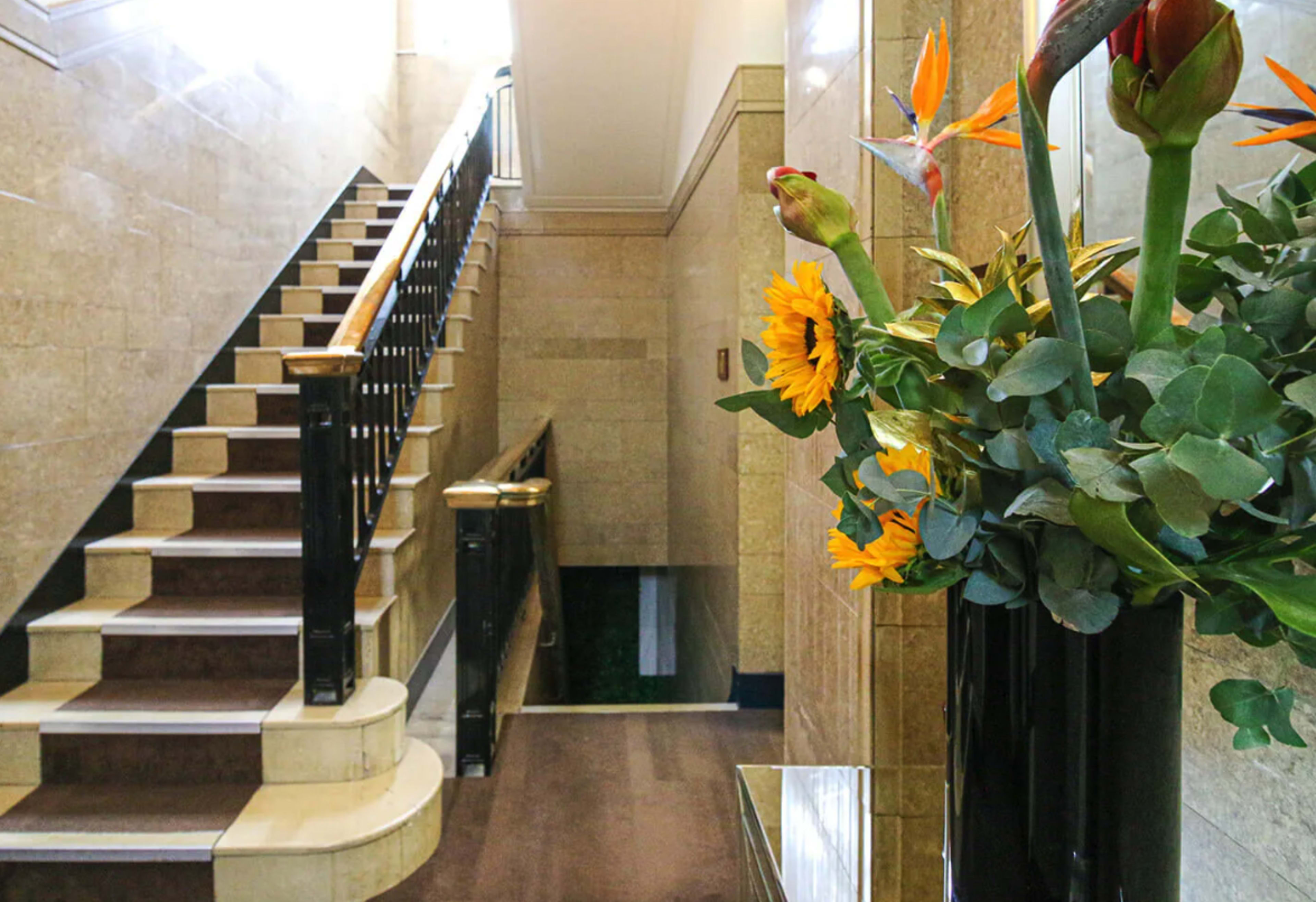 The image shows a well-lit staircase with a polished wooden handrail, flanked by a large vase of flowers beside a wall.