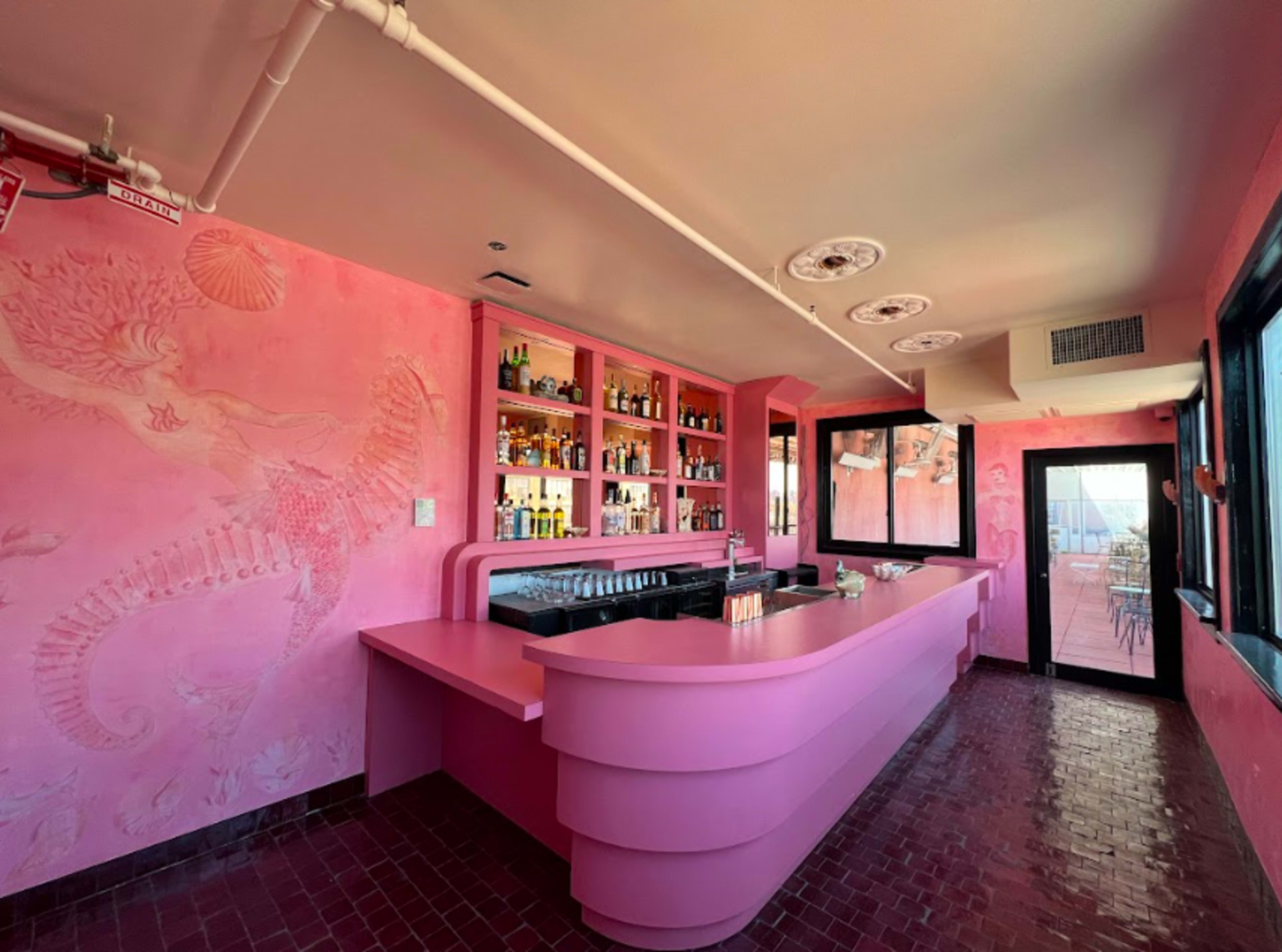 The image shows a pink-themed bar with a curved counter, shelves stocked with various bottles, and large windows allowing natural light to enter.