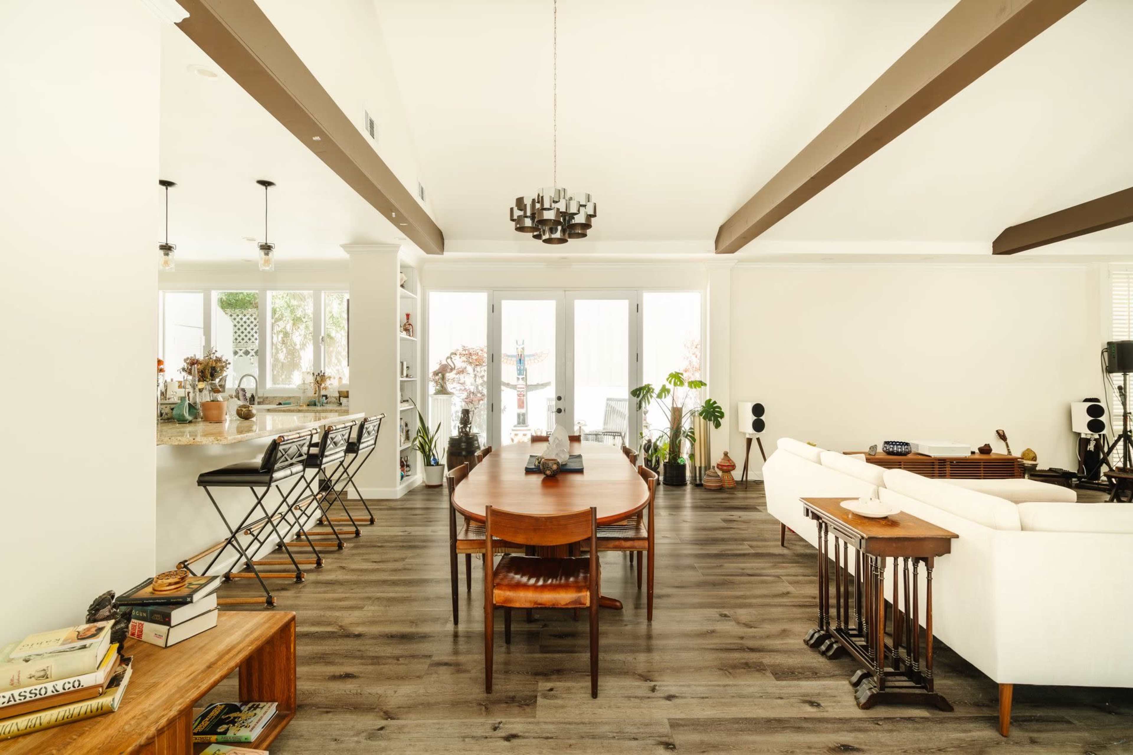 The image shows a spacious interior with a dining area featuring a round table surrounded by chairs, a modern kitchen with bar seating, and a comfortable living space furnished with a couch and various decorative plants.