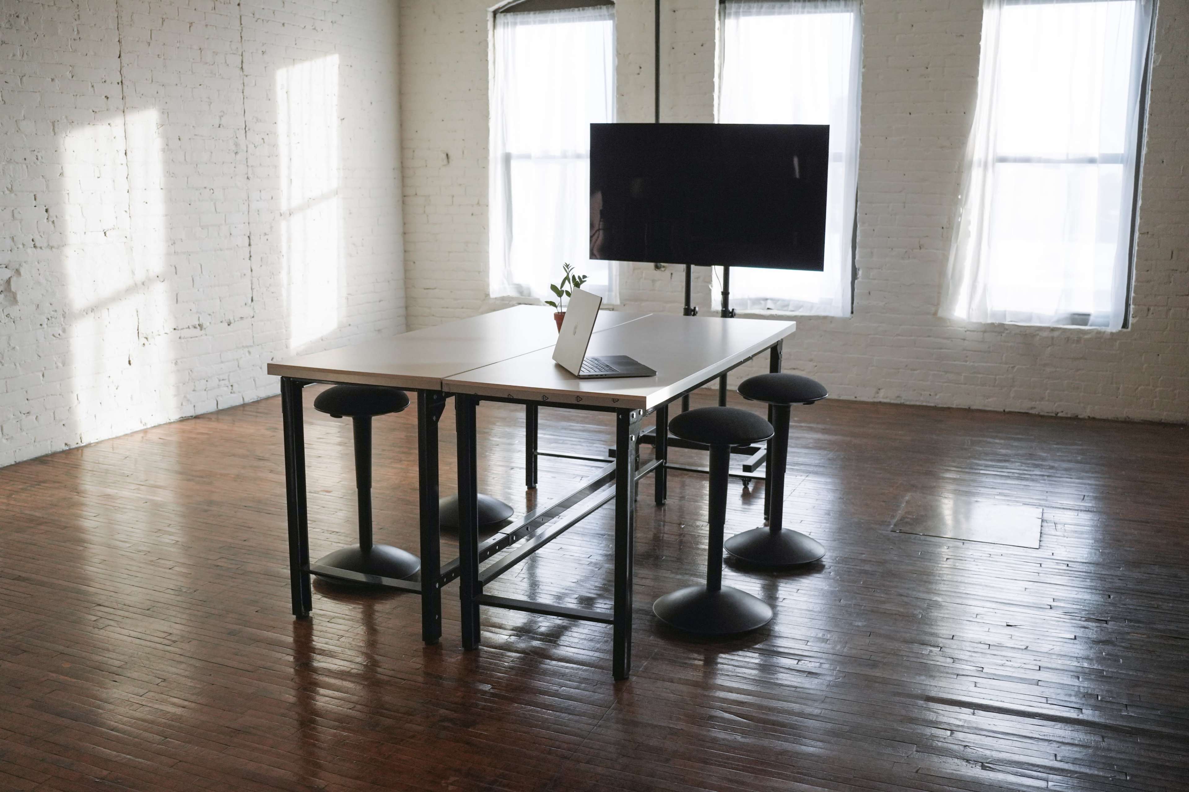A minimalist workspace features a table with a laptop and a potted plant, surrounded by four stools, in a well-lit room with large windows.
