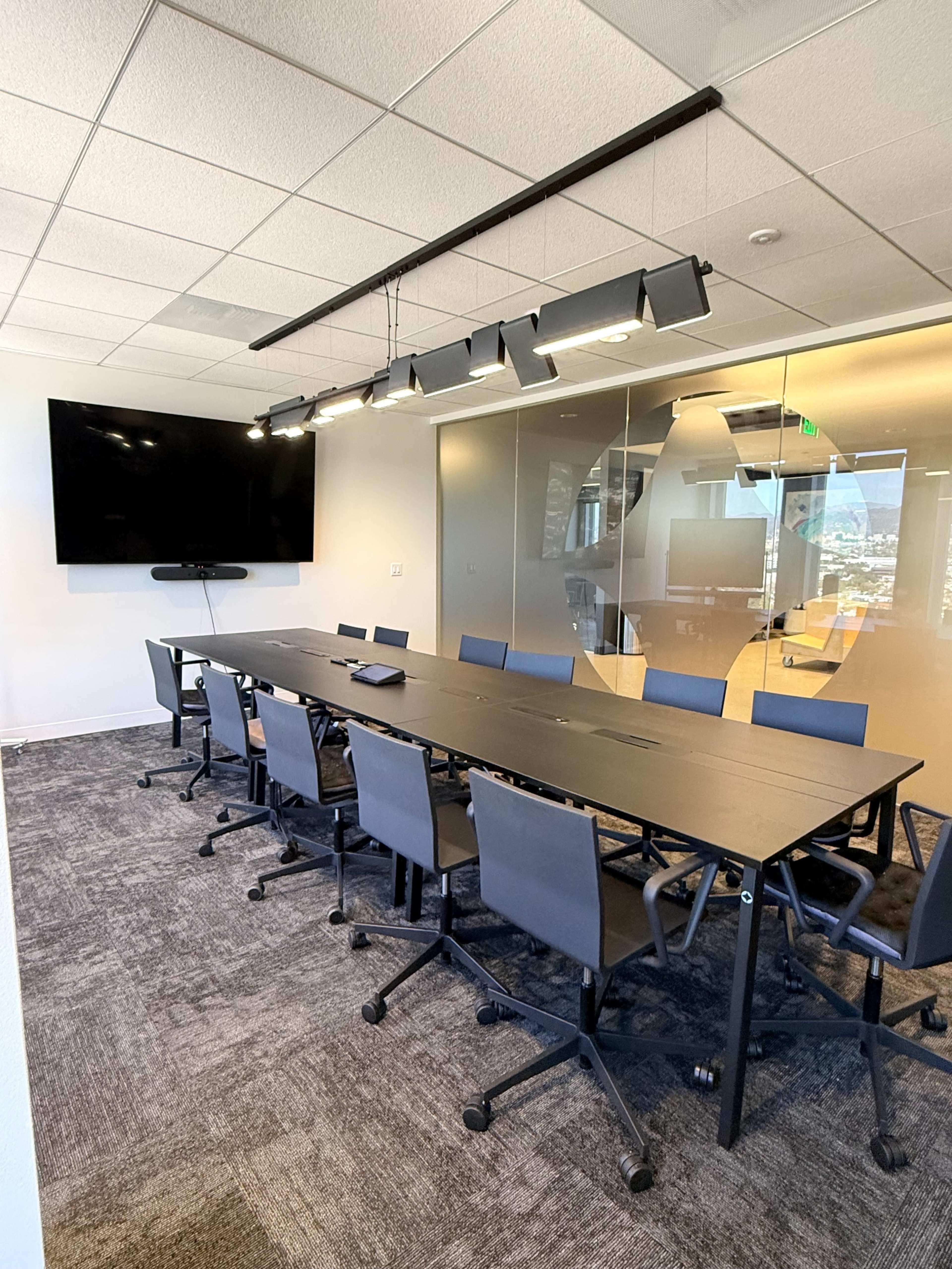 A modern conference room features a long black table surrounded by rolling chairs, with a large screen mounted on the wall and ample natural light from large windows.