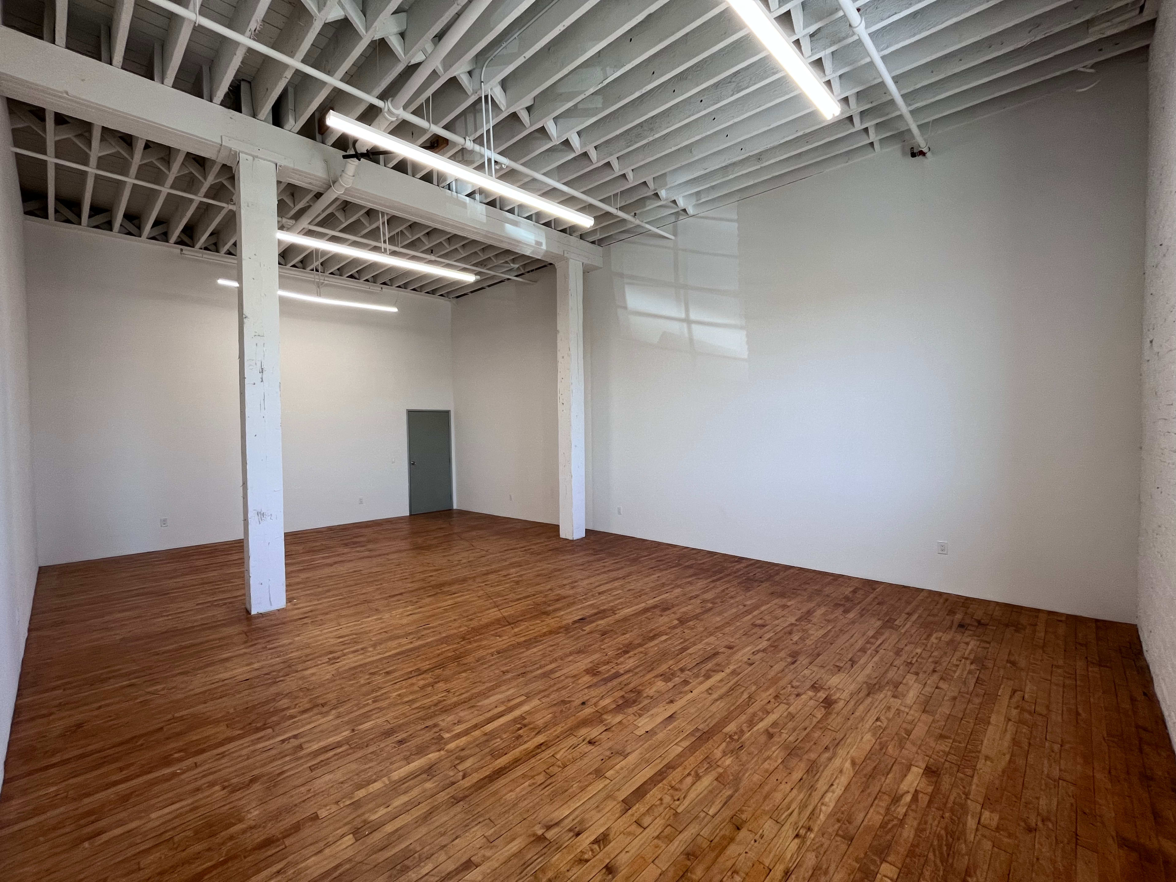 An empty room with a wooden floor, white walls, exposed beams, and overhead lighting.