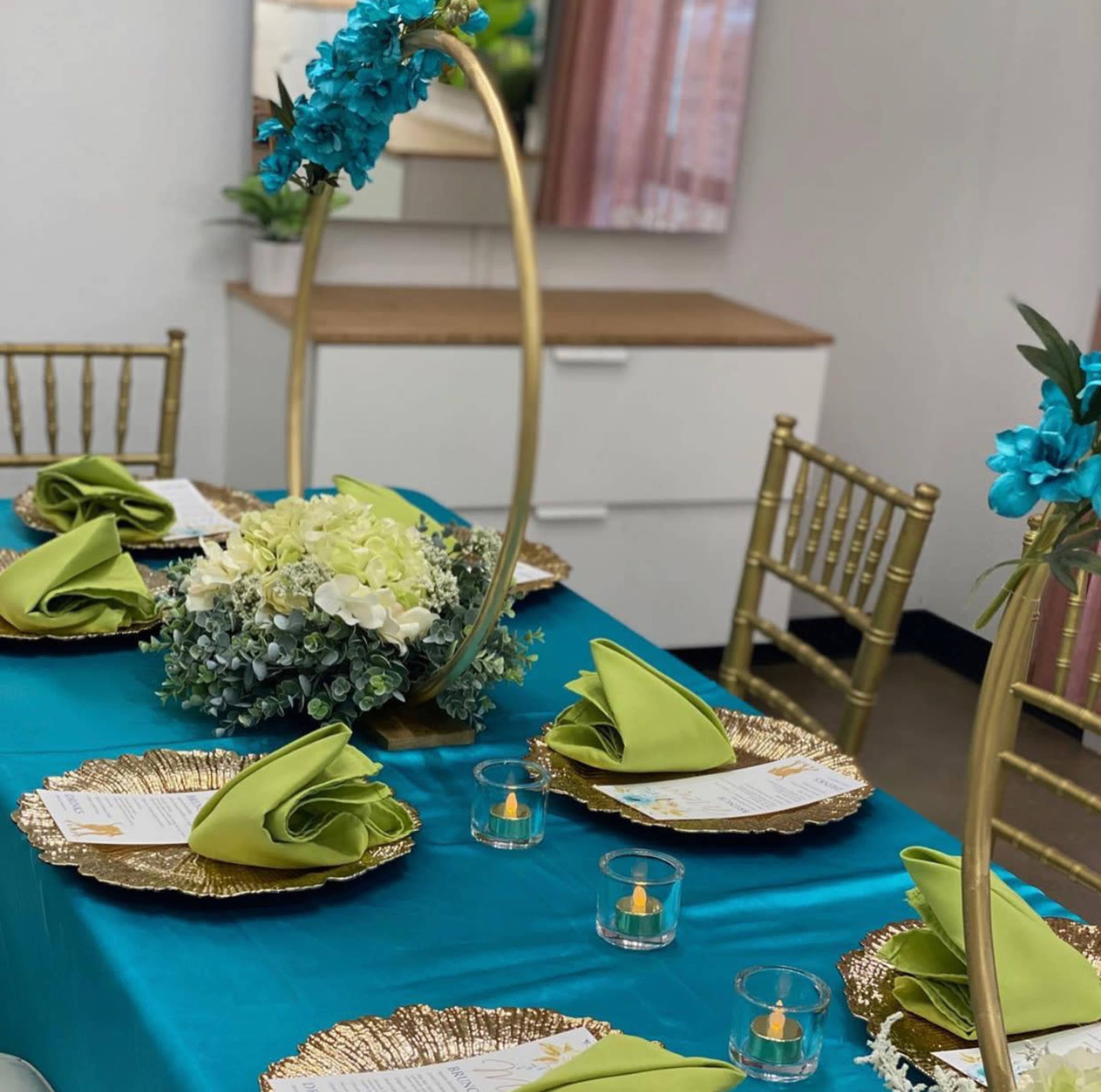 A neatly arranged dining table features gold chargers, green napkins, and a floral centerpiece, with gold chiavari chairs surrounding the setup.
