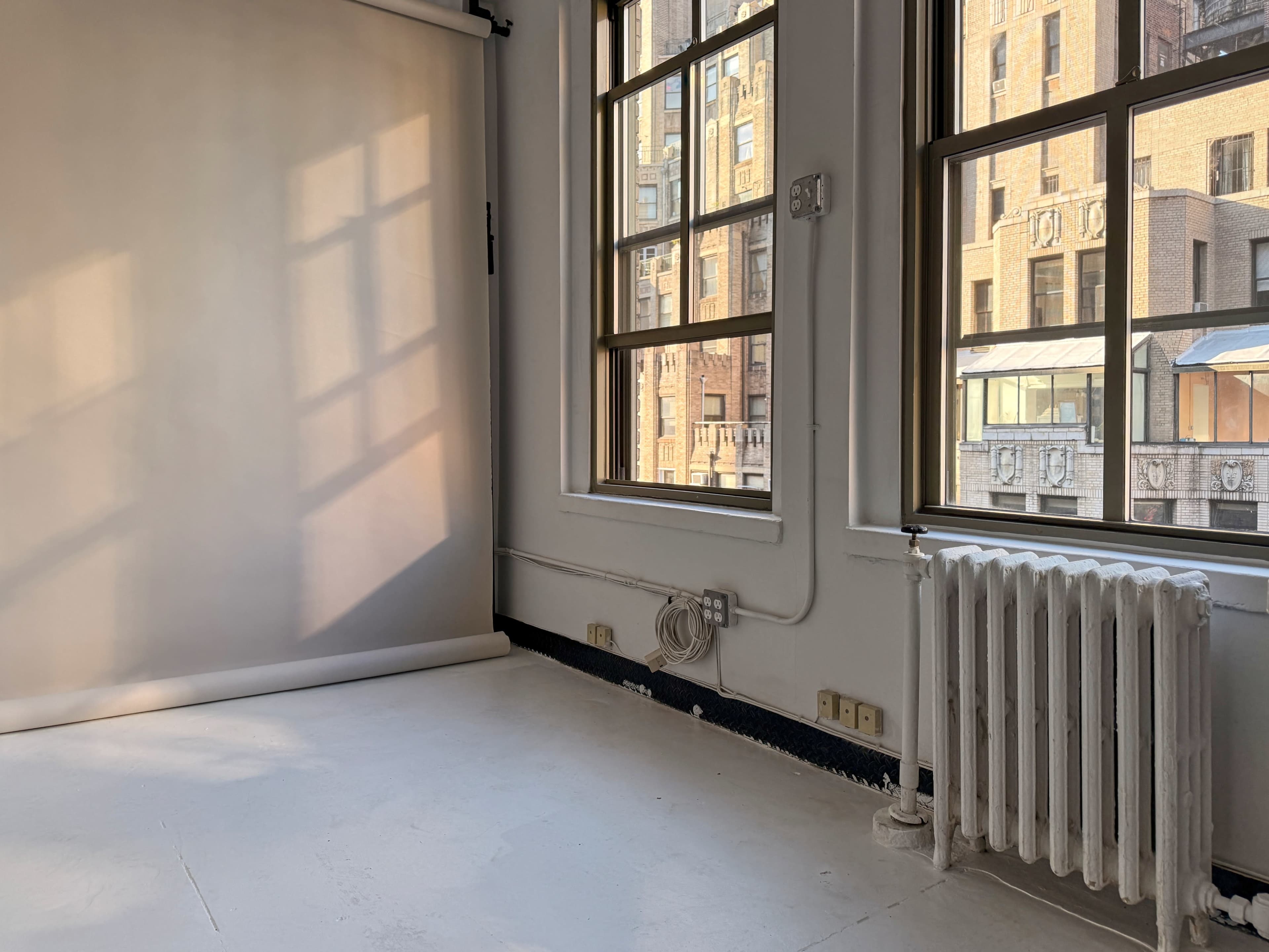 A bright, empty room features large windows with cityscape views, a backdrop stand, and a white radiator along the wall.