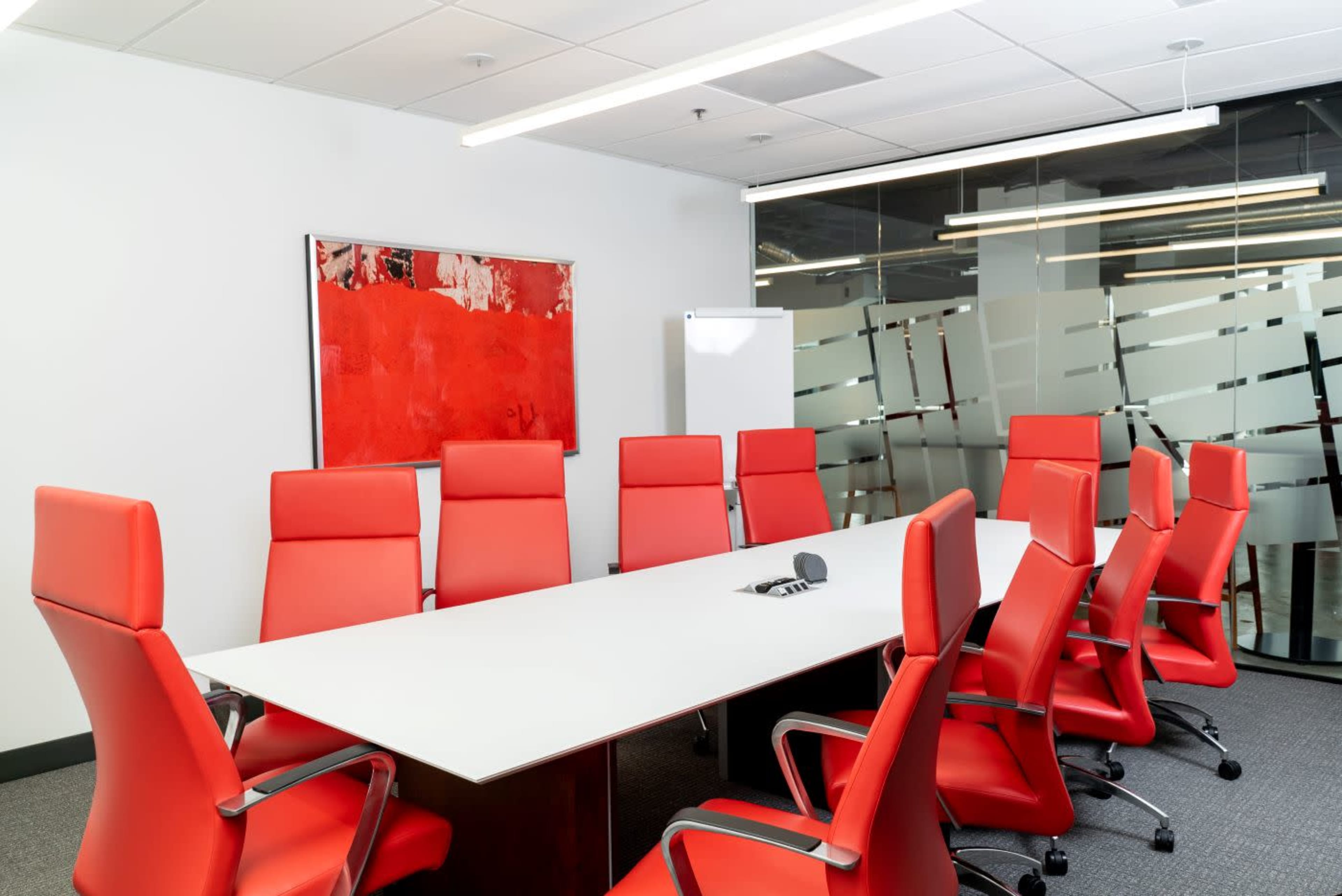A modern conference room features a long white table surrounded by eight red chairs, with a large red artwork on the wall and glass partitions.