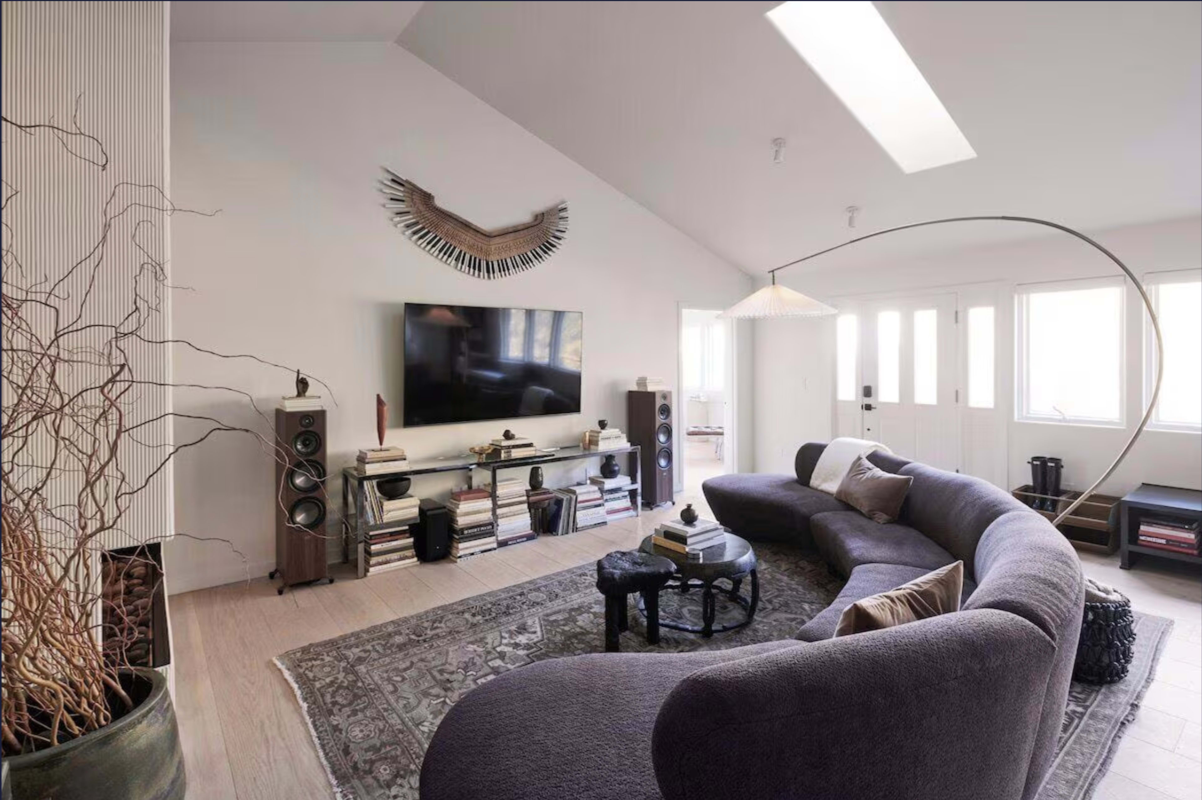 A modern living room features a curved sofa, a large television on a low shelf, and natural light streaming through windows.