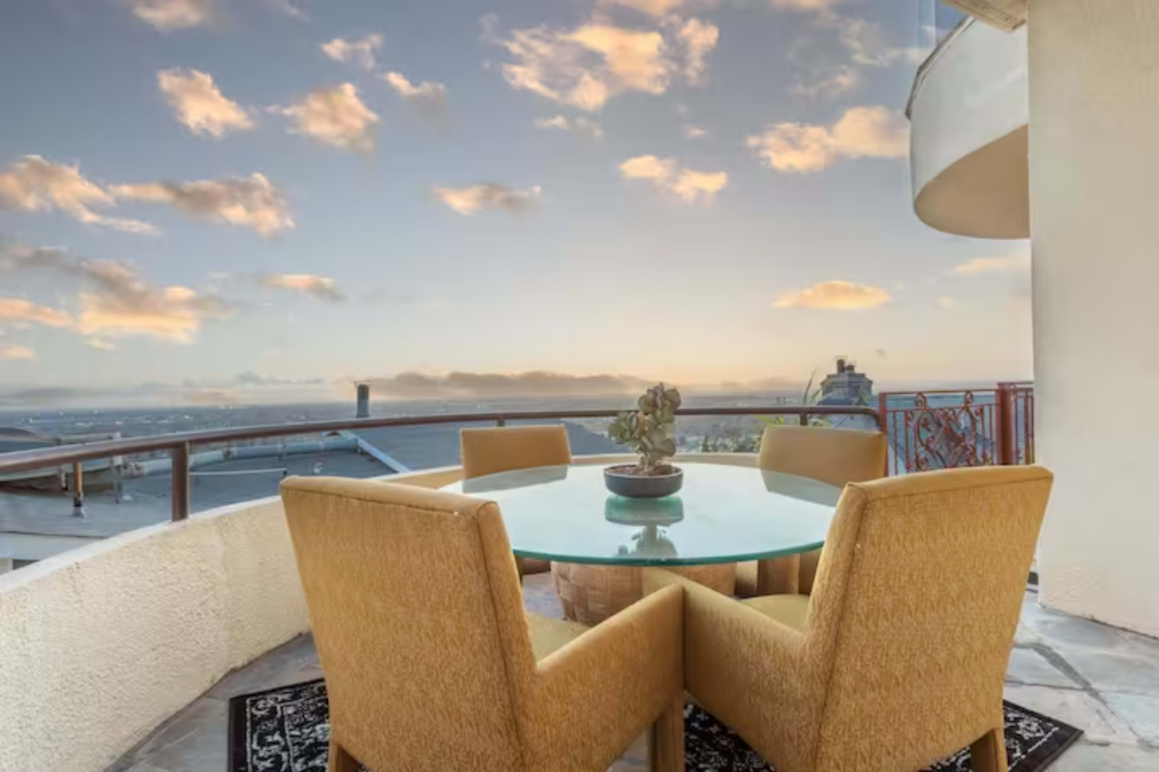 A circular glass table with four yellow chairs is set on a balcony overlooking a sunset sky and distant rooftops.