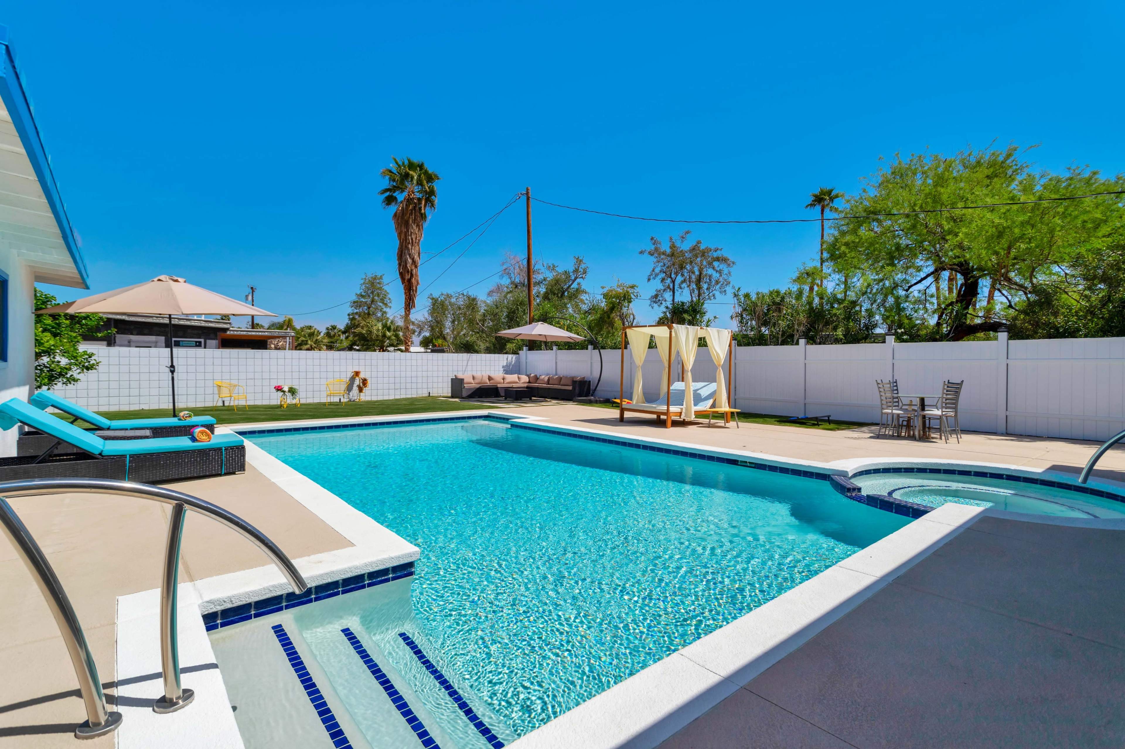 A backyard pool area featuring a large swimming pool, a hot tub, lounge chairs, and a shaded seating area surrounded by palm trees and greenery.