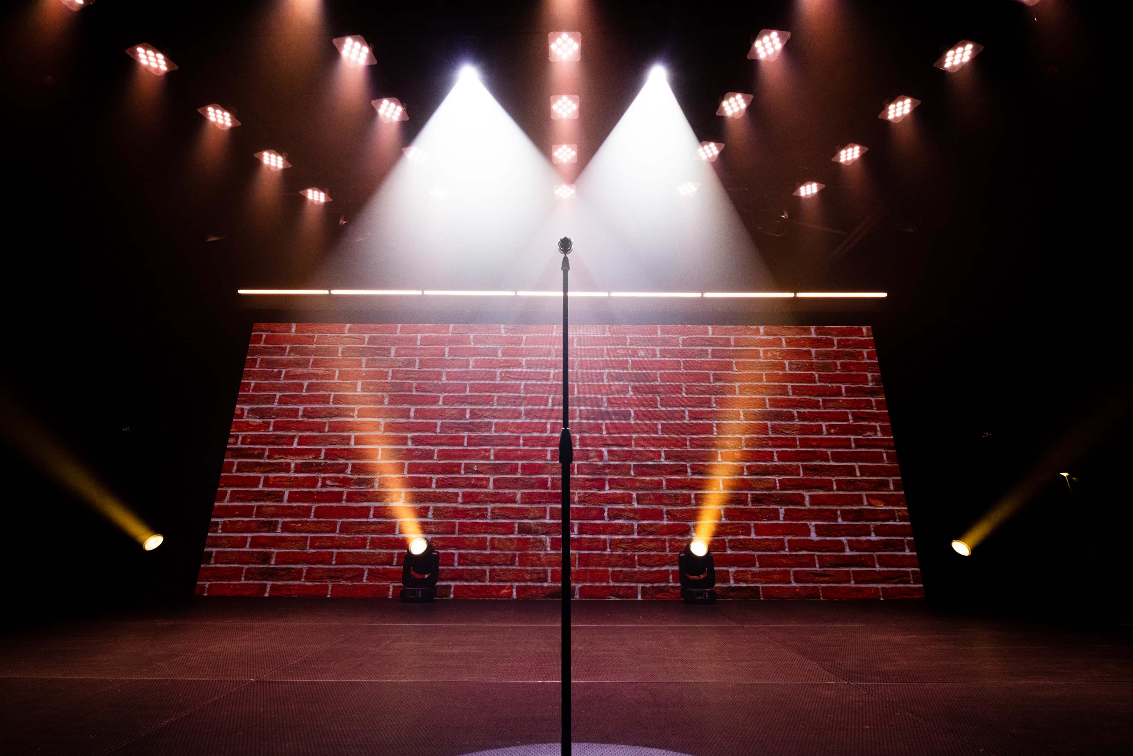 A microphone stand is centered on a stage with a brick wall backdrop and bright lights shining down.