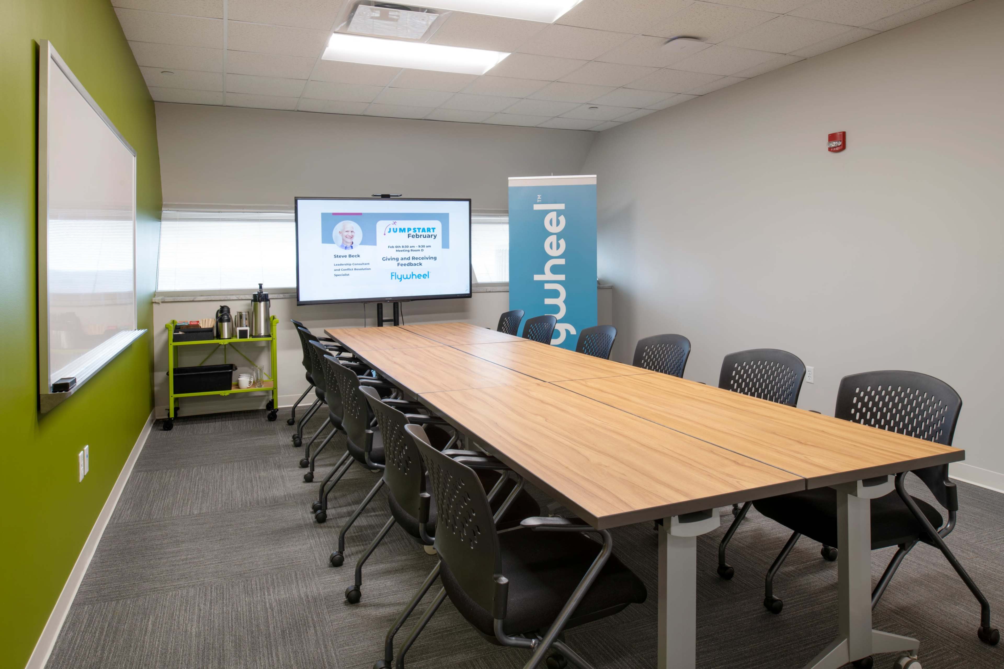 A modern conference room with a long wooden table, office chairs, a whiteboard, and a screen displaying a presentation.