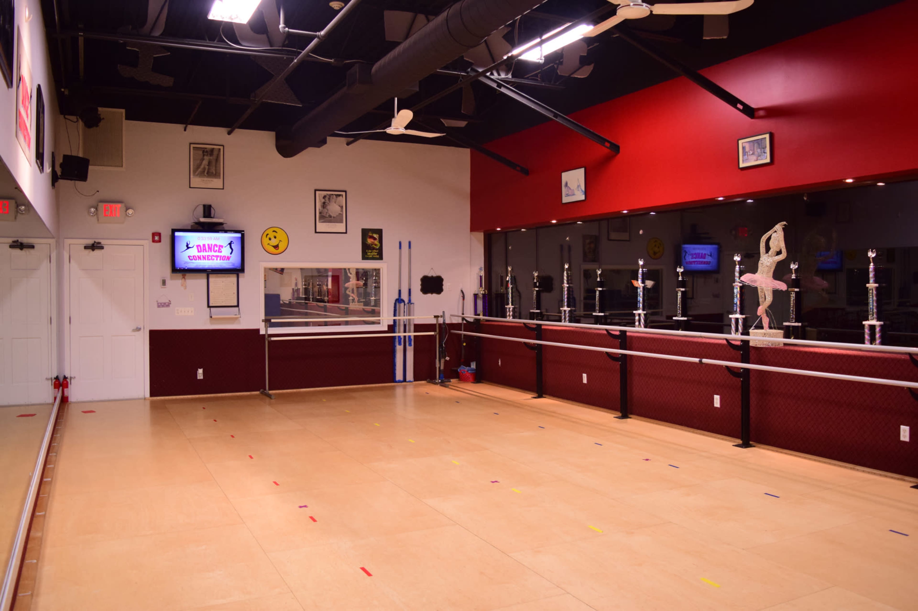 A spacious dance studio with a wooden floor, mirrored walls, and a barre along one side, featuring various trophies and a television display on the wall.
