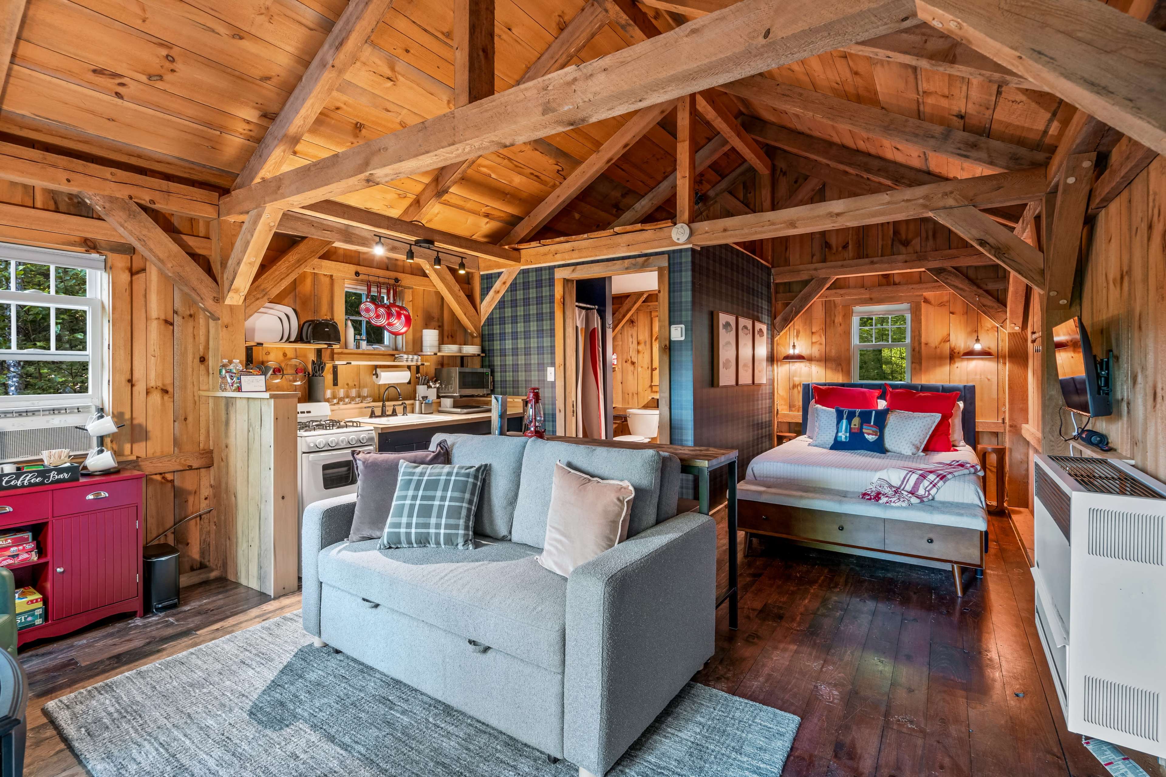 The image shows a cozy cabin interior featuring wooden beams, a small kitchen area, a seating space with a gray couch, and a double bed with colorful pillows.