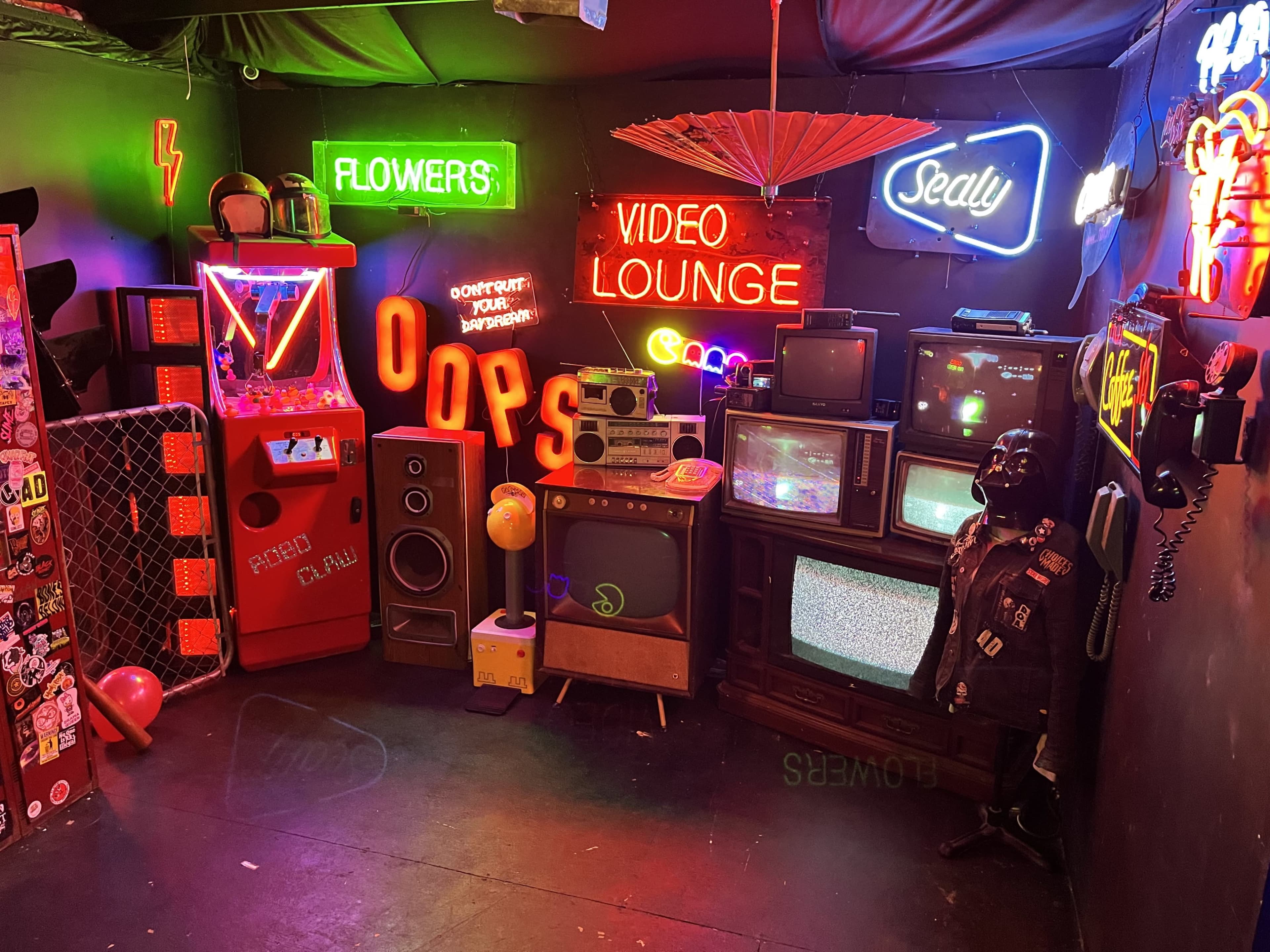 The scene features a dimly lit lounge adorned with various vintage televisions, neon signs, a pinball machine, and eclectic decorations.