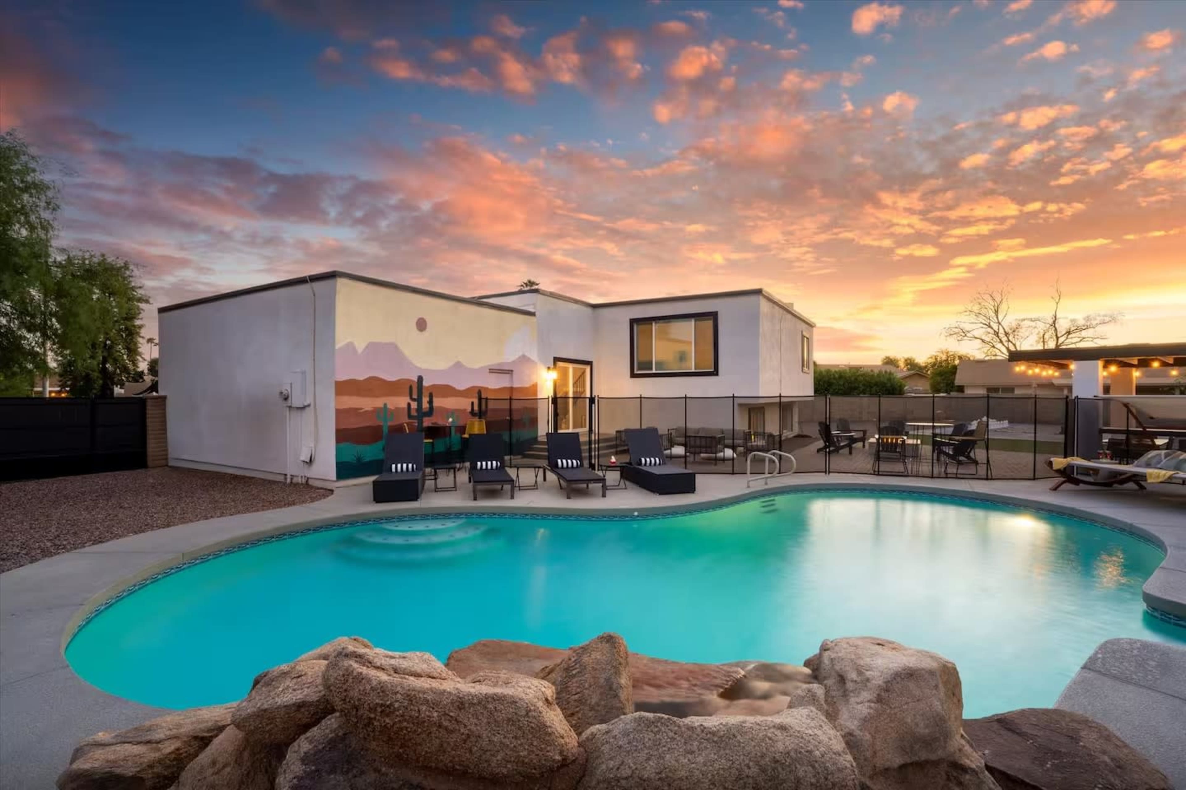 A swimming pool with rock features sits in front of a modern home with a mural, under a colorful sunset sky.