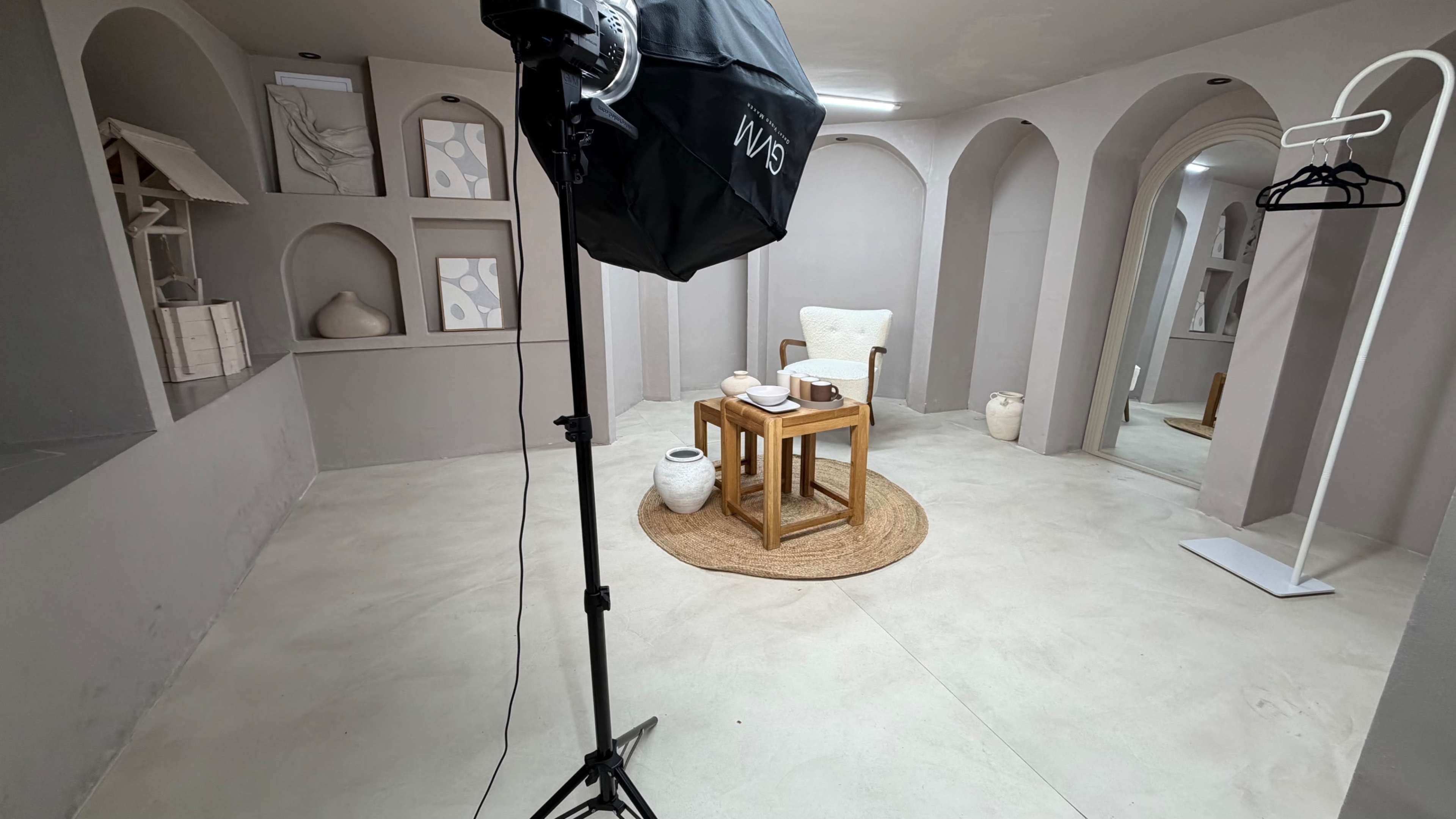 The image shows a minimalist studio space with a wooden table and a white chair, surrounded by light-colored walls and decorative items in alcoves.