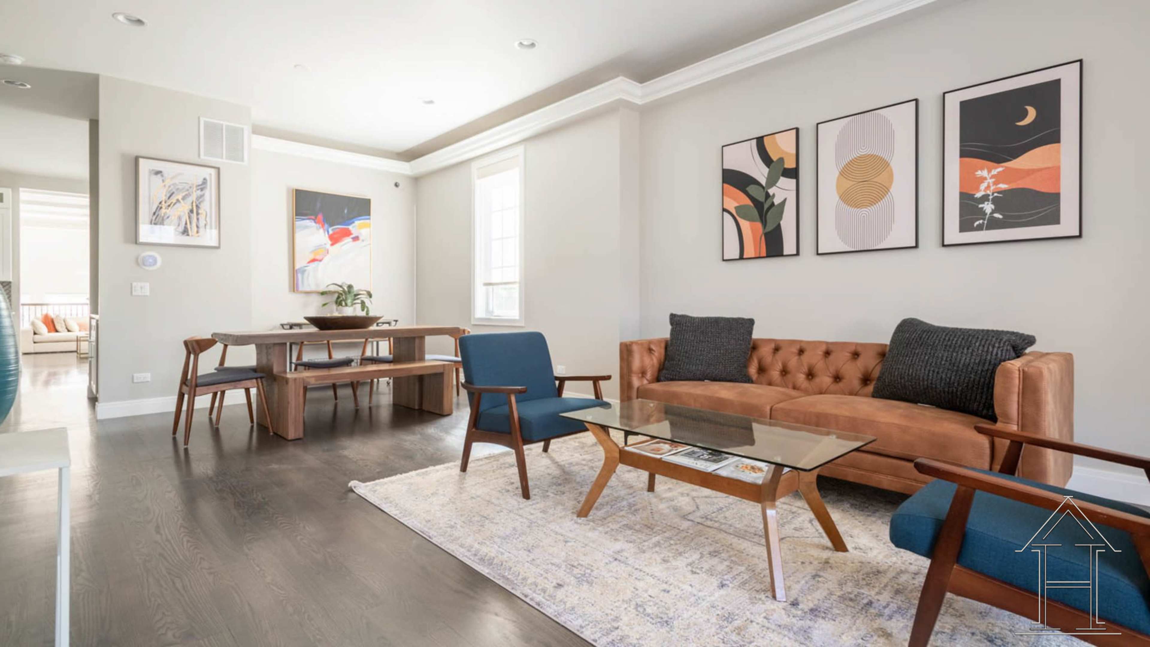 A modern living room with a brown tufted sofa, blue chairs, a glass coffee table, and framed artwork on the walls.