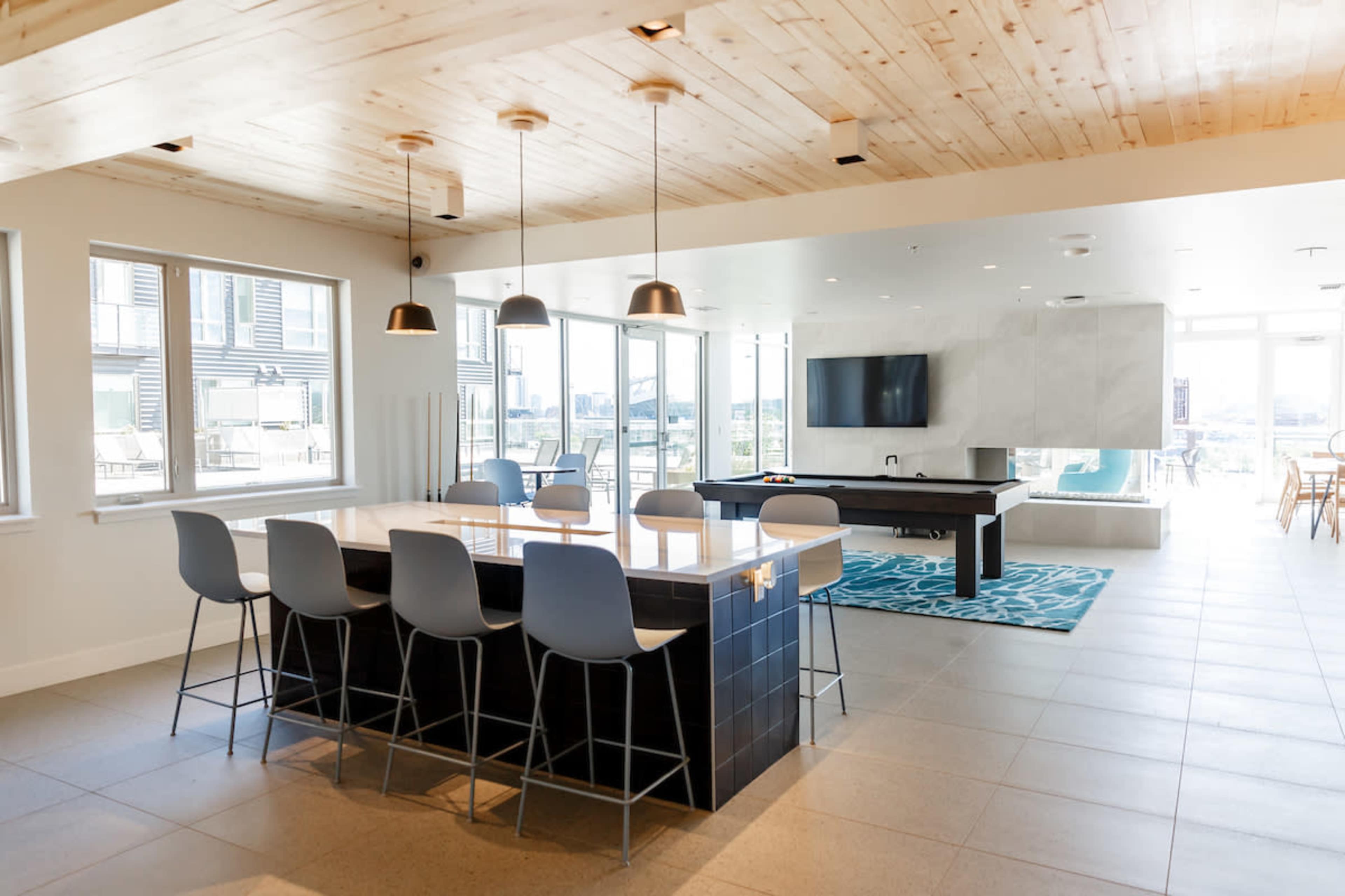 A modern open-concept lounge area with a kitchen island, seating, and a pool table, illuminated by pendant lights and large windows.