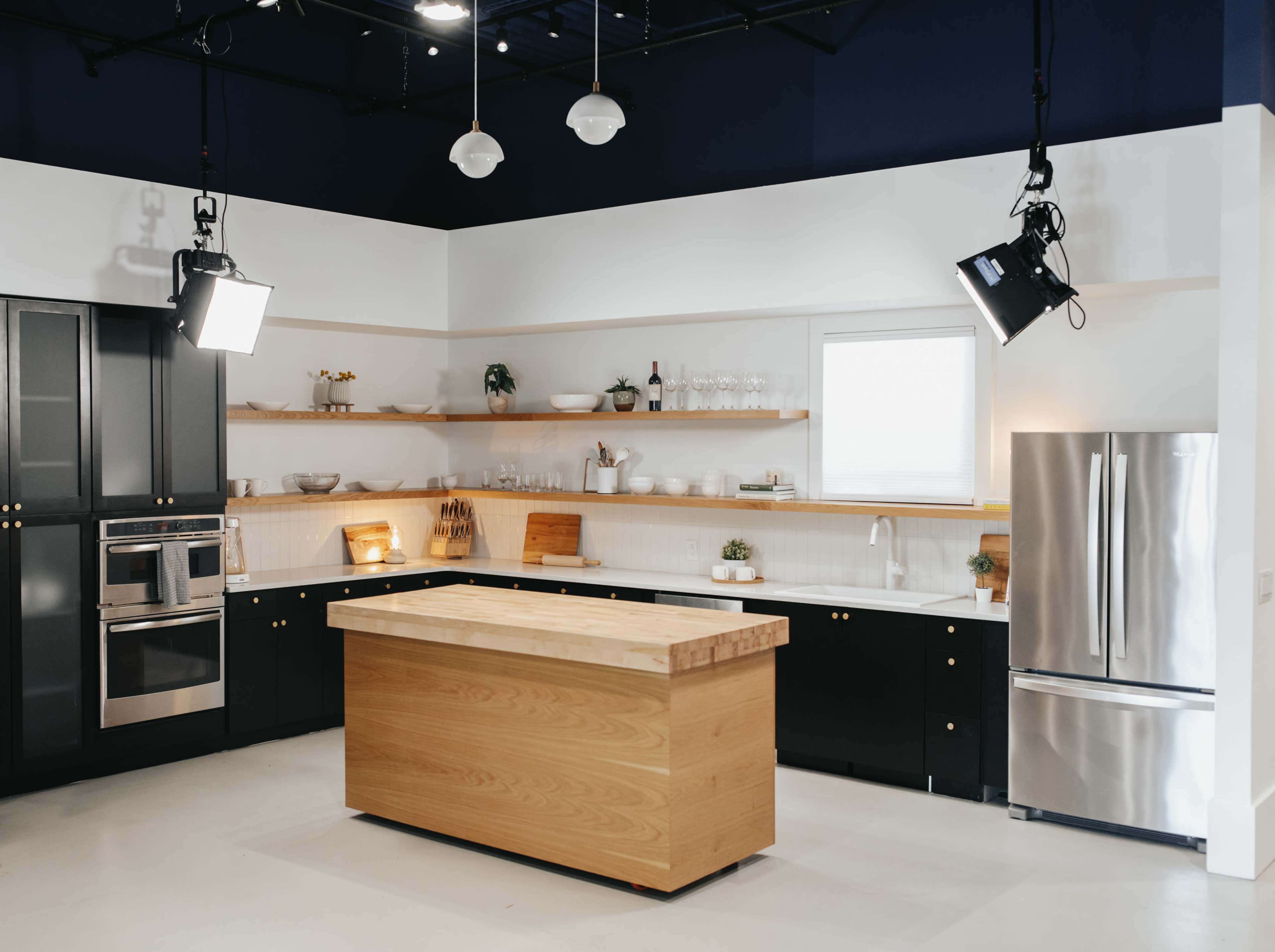 The image shows a modern kitchen with black cabinetry, a wooden island, and stainless steel appliances, featuring open shelving with kitchenware.