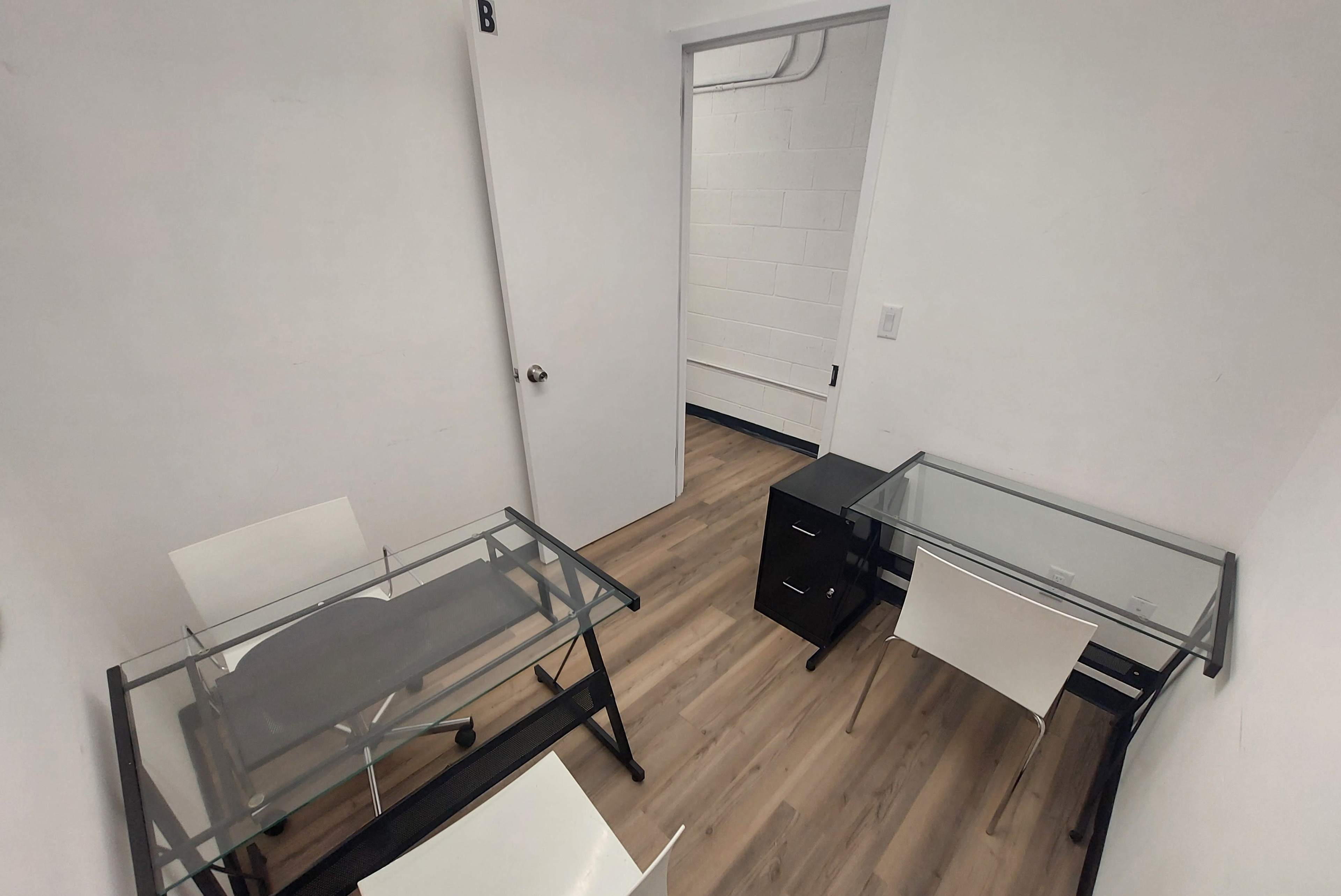 The image shows a small office space with two glass-top desks, black office chairs, and a filing cabinet near an open door.