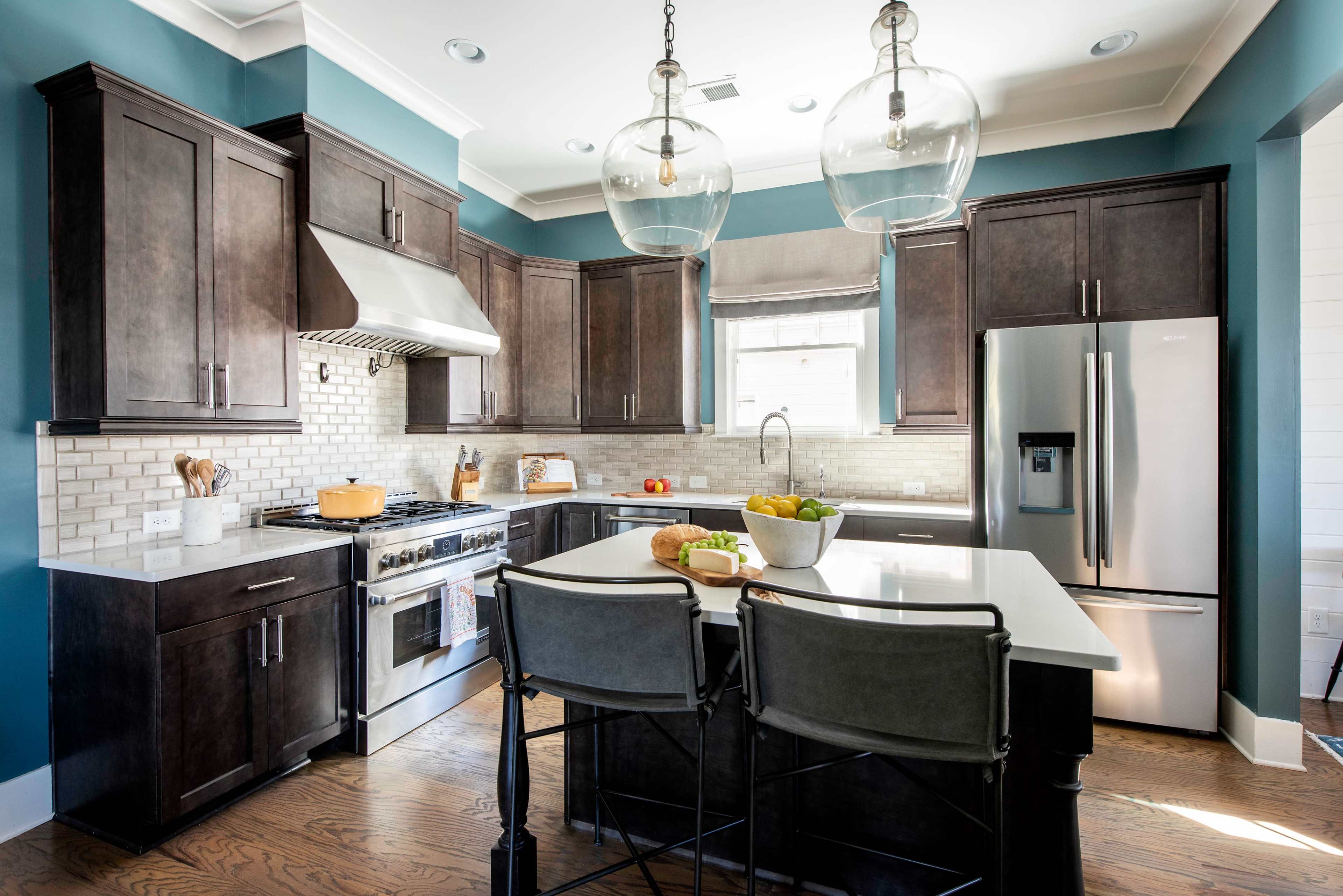 The image shows a modern kitchen with dark cabinets, a large island with bar stools, stainless steel appliances, and pendant lighting.
